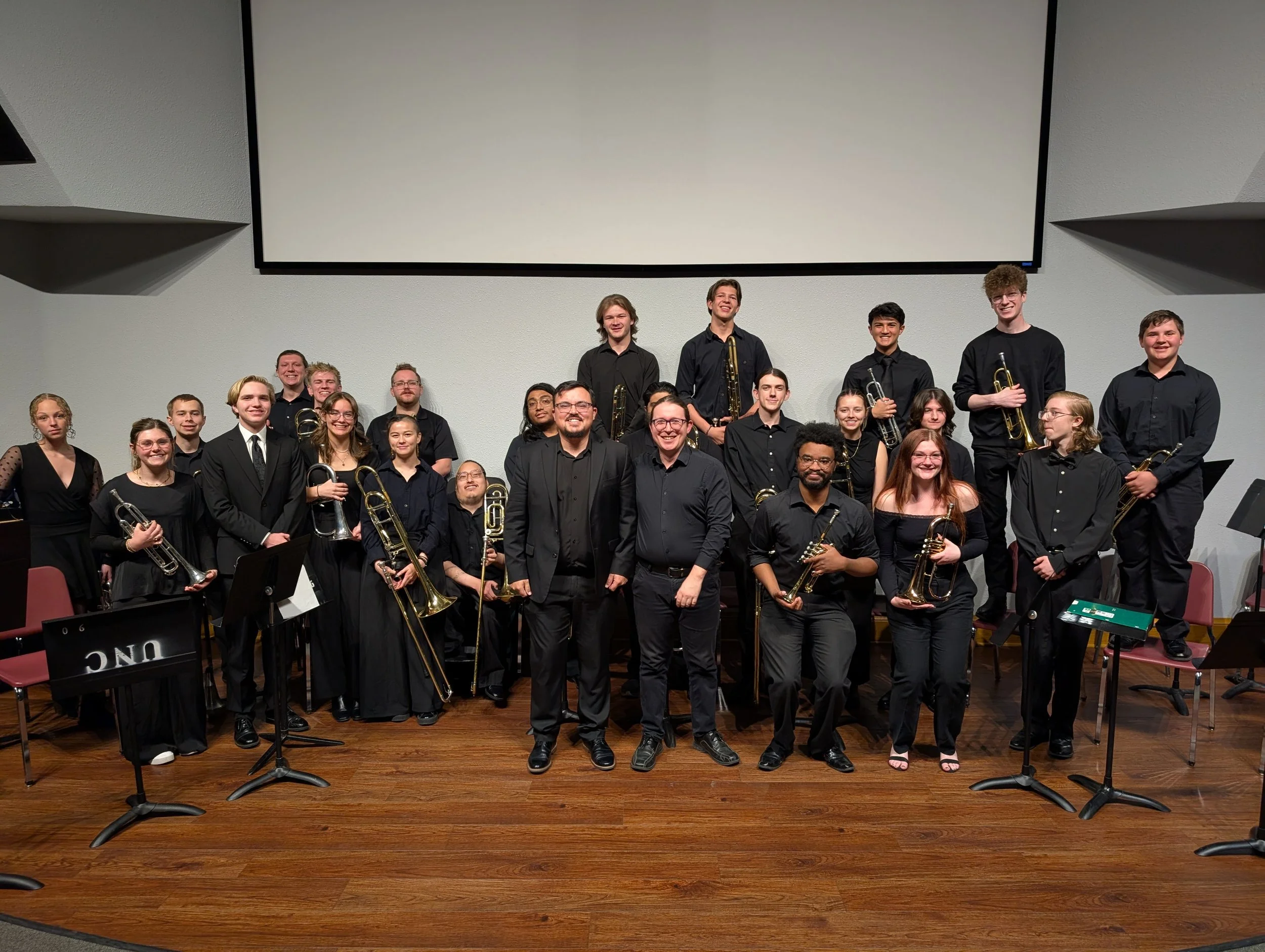 A group of 21 musicians, mostly young adults, dressed in black, standing on stage with their instruments, posing for a photo after a performance or rehearsal.