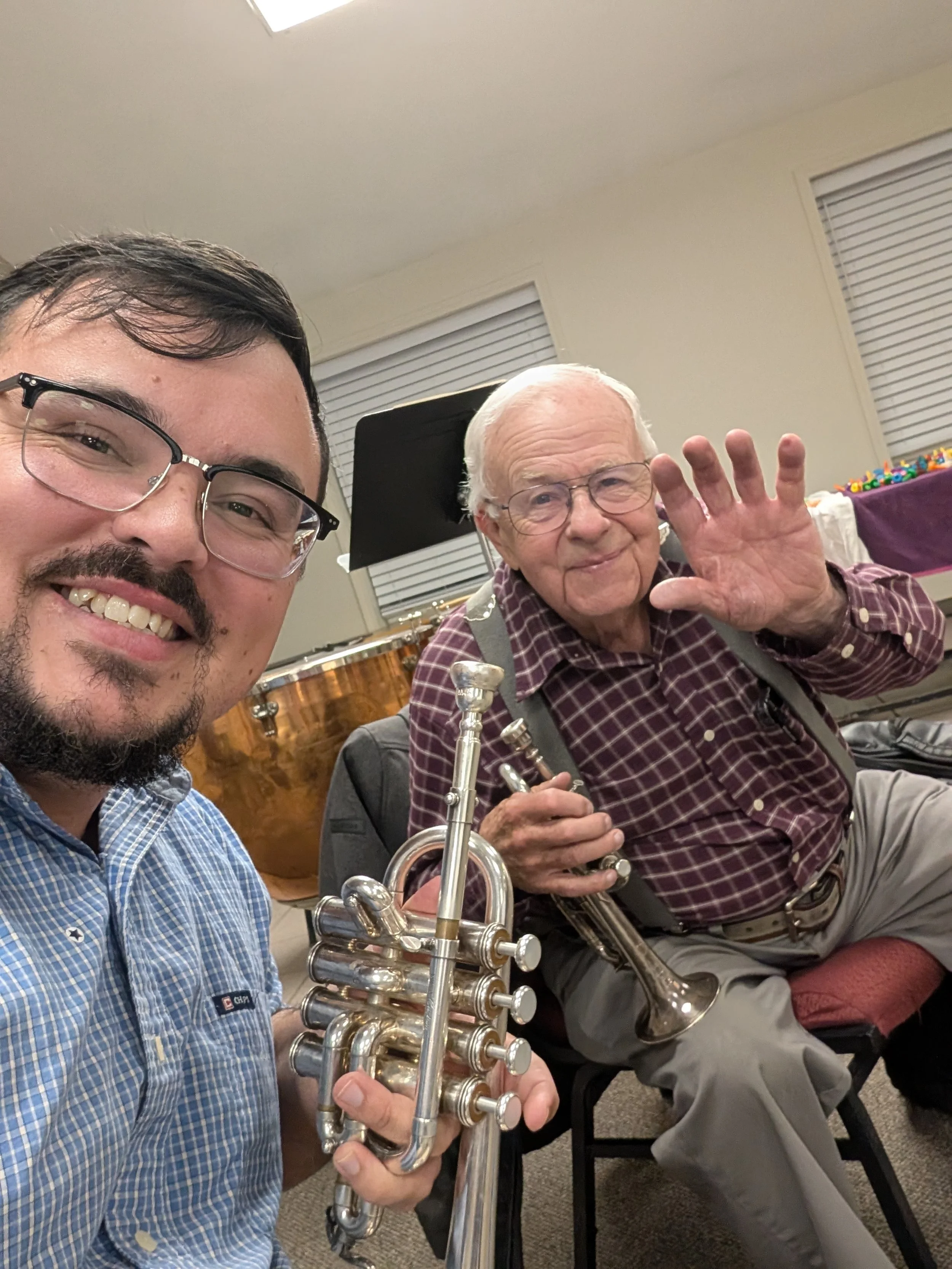 Two men smiling, one young with glasses holding a trumpet, and an elderly man waving, in a room with musical instruments and a table with music accessories.