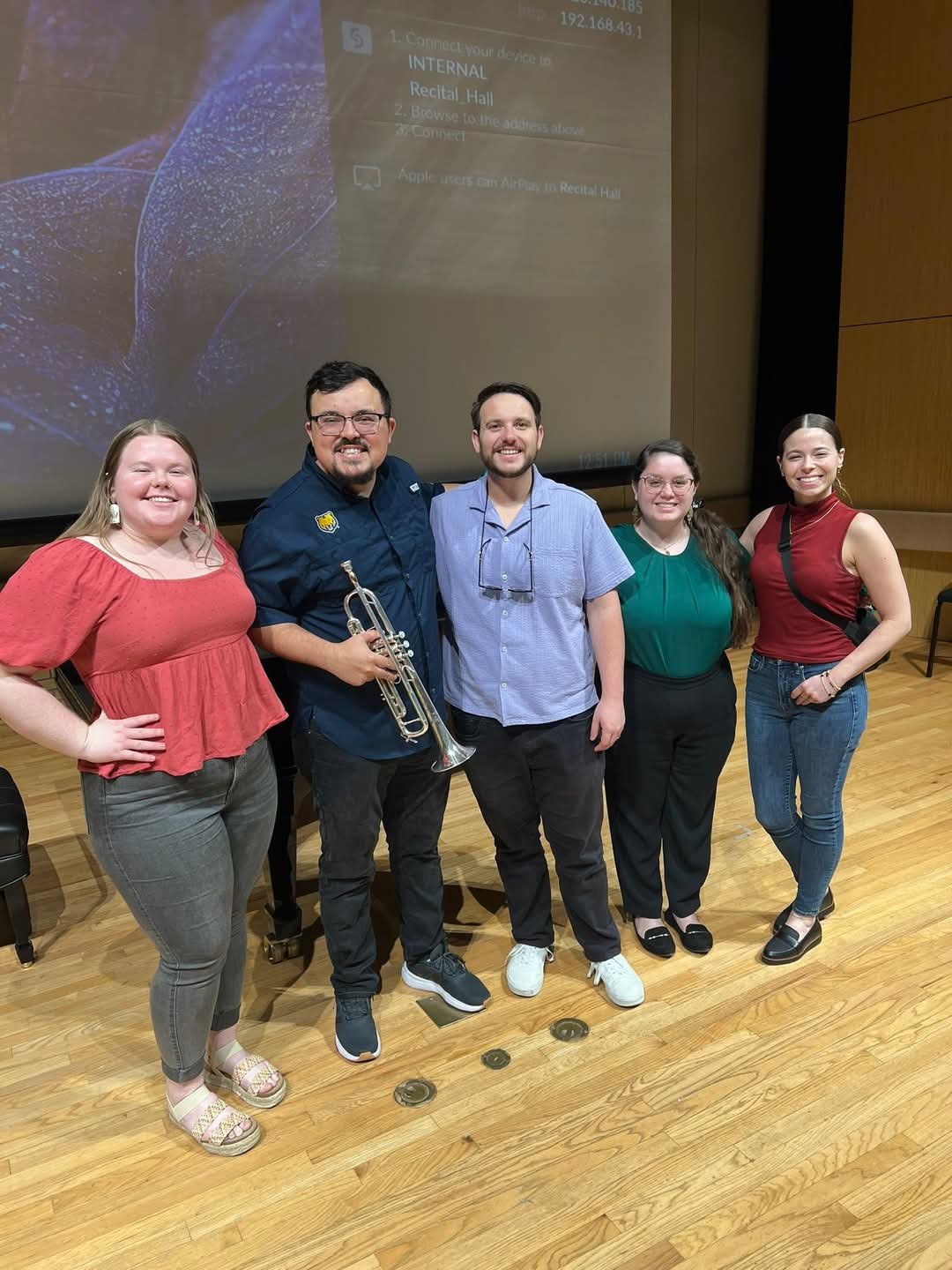 Group of five young adults standing on a wooden floor in a conference room, posing for a photo. They are smiling and dressed casually, with one person holding a trumpet. There is a large projection screen behind them with some text visible.