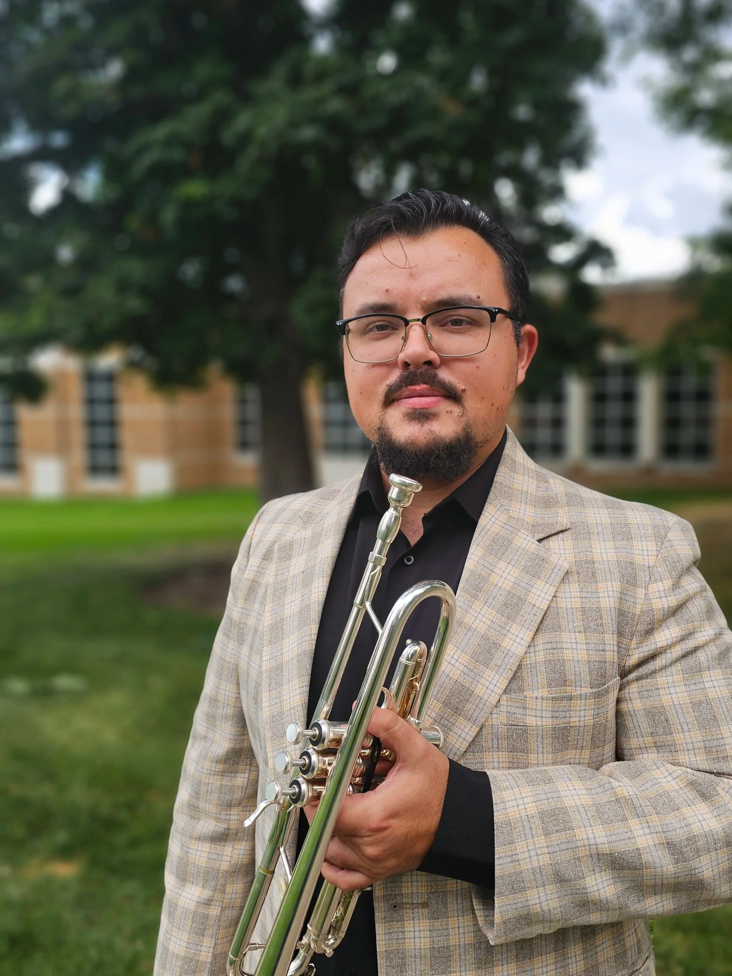 A man with dark hair and glasses holds a trumpet outdoors with a background of trees and a building.