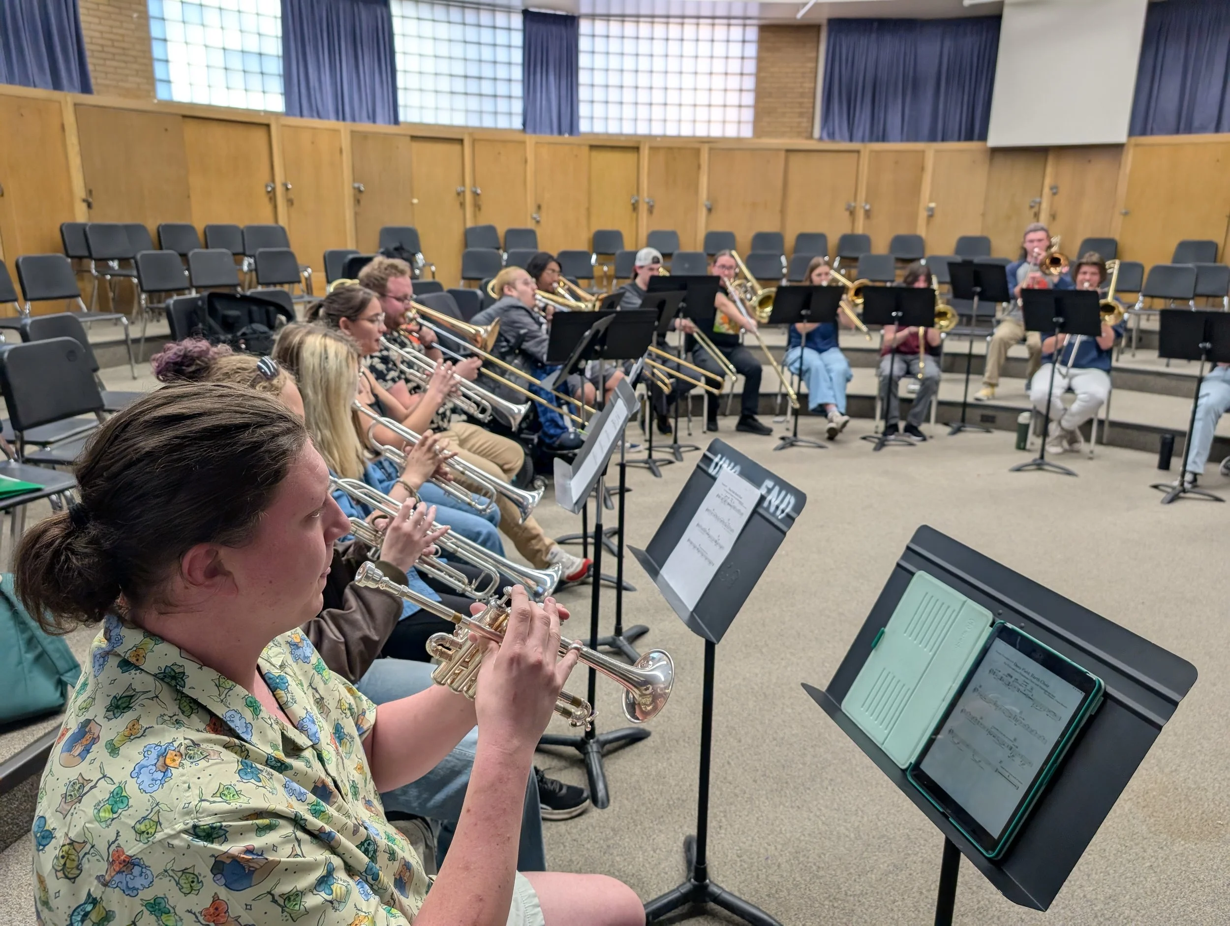 Group of young musicians playing brass instruments during a rehearsal in a music room with chairs and music stands.