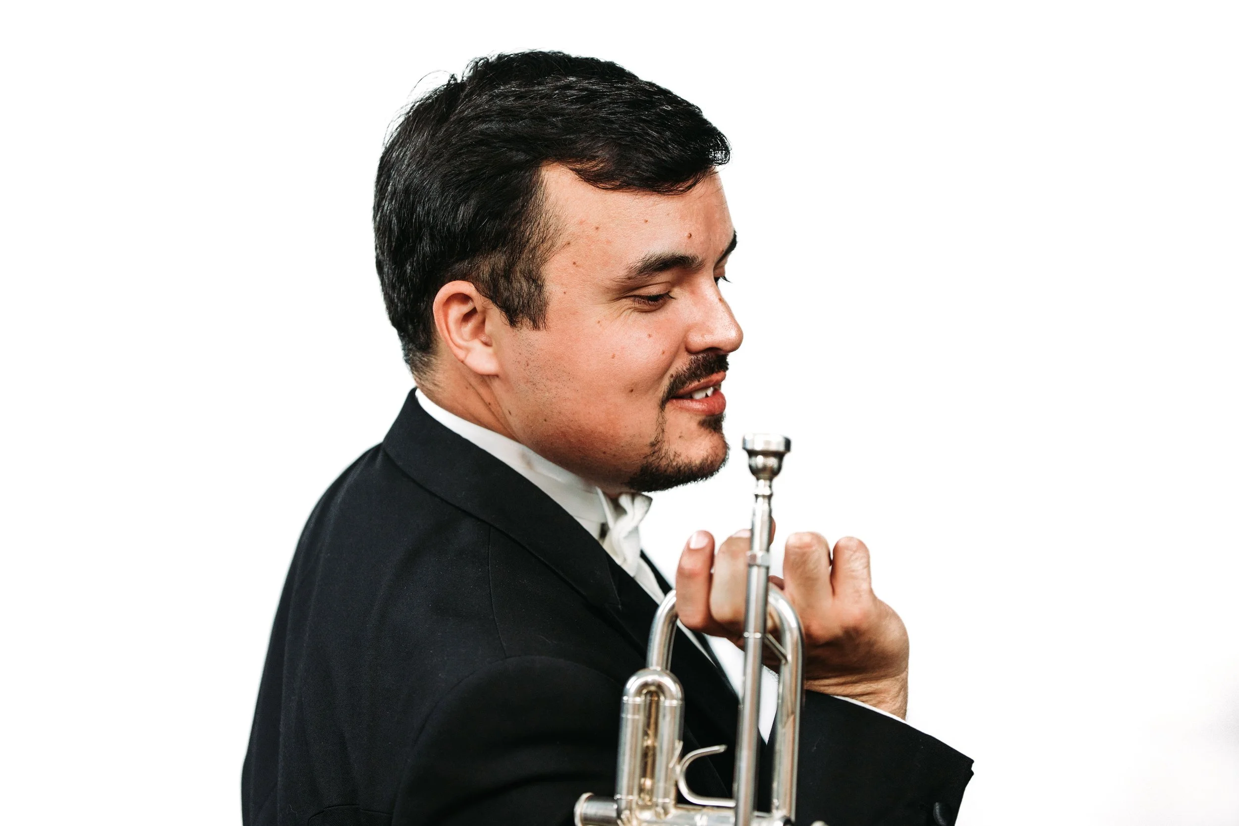 A man in a tuxedo holding a trumpet close to his shoulder, smiling and looking down, against a plain white background.
