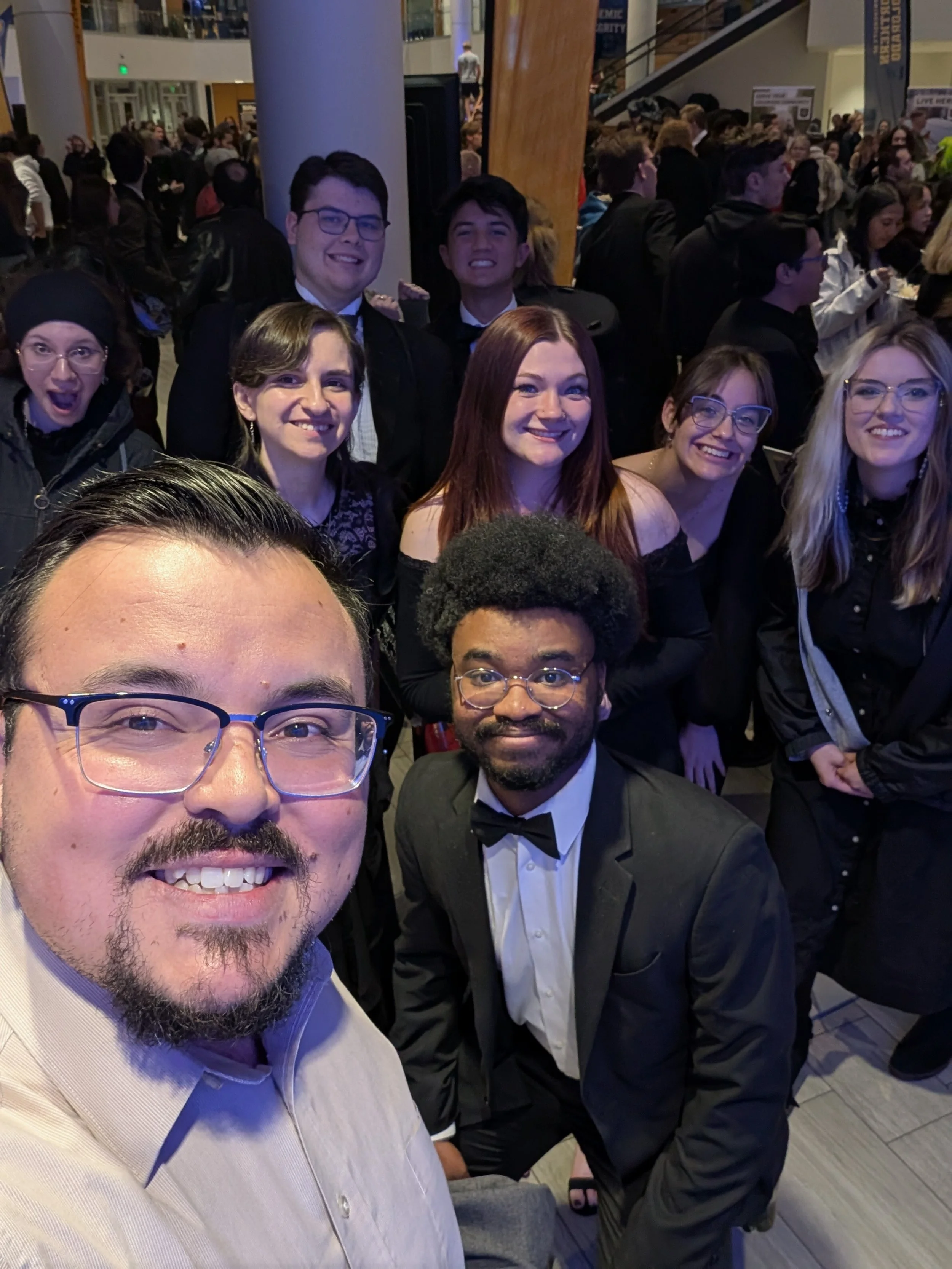 Group of diverse young adults dressed in formal attire, taking a selfie at a crowded event, some smiling, in an indoor venue with a wooden pillar in the background.