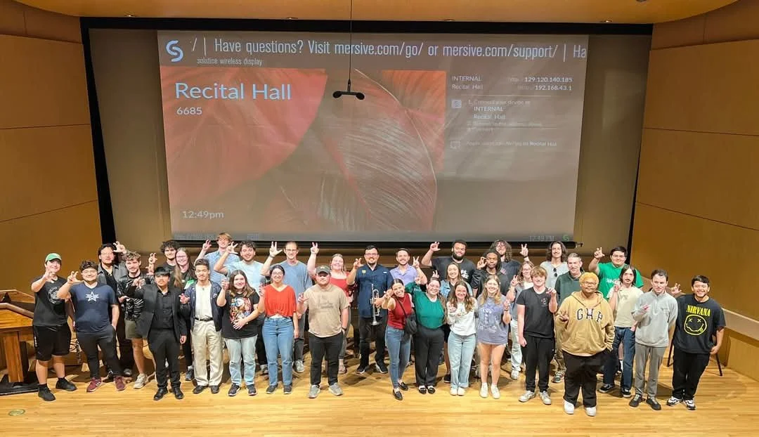 Group of people standing on stage in front of a large screen, all making peace signs and smiling.