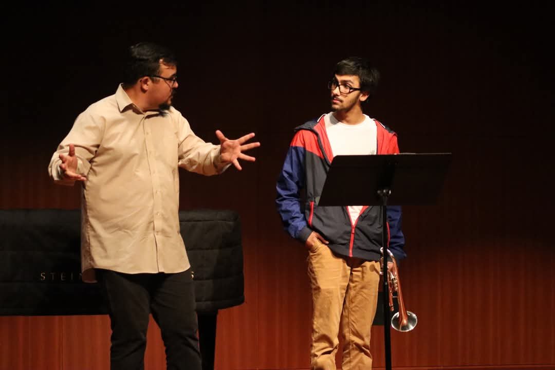 Two men engaged in a conversation on a stage, one gesturing with hands, the other with hands in pockets, with a music stand and a piano in the background.