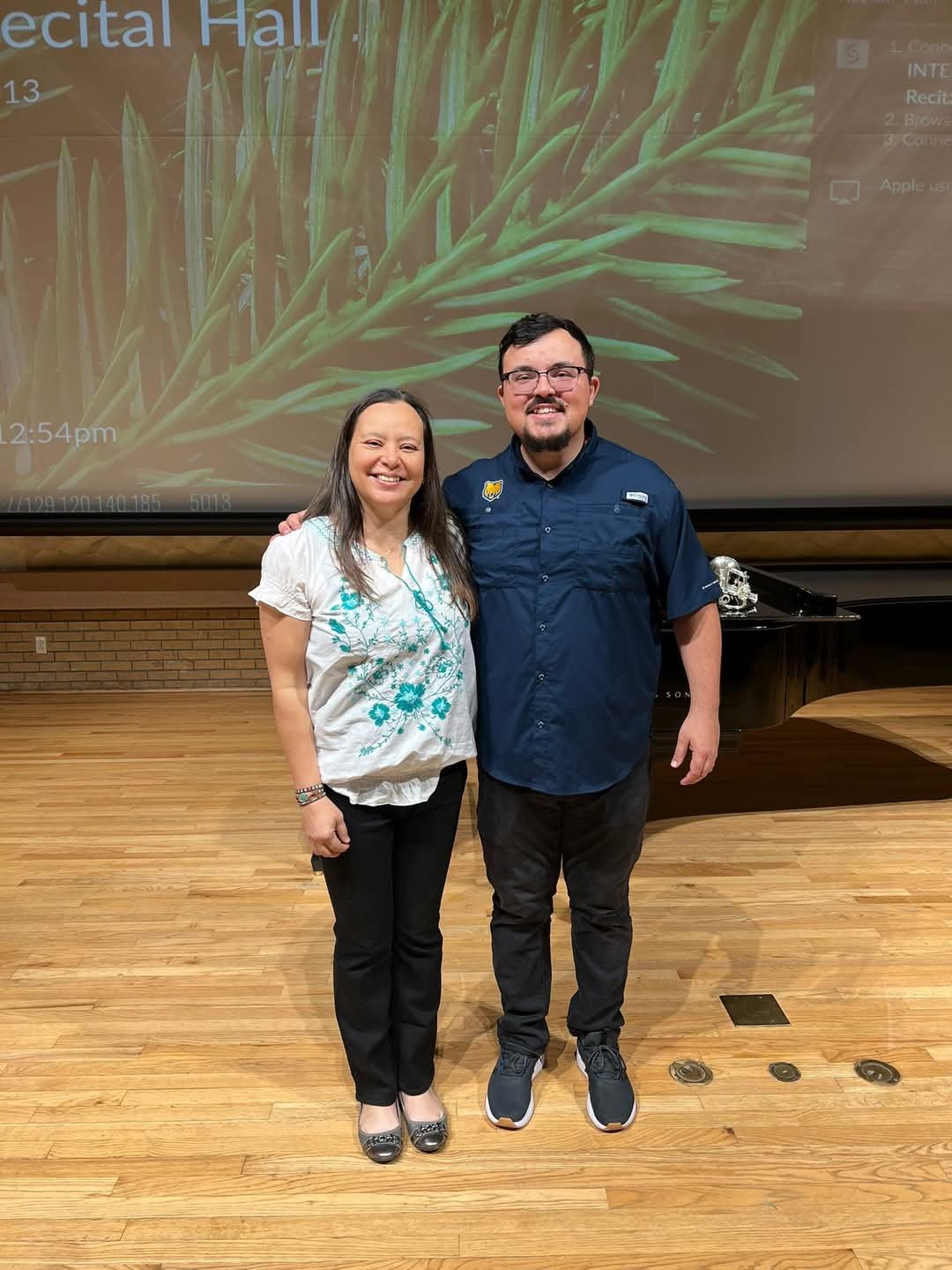 A woman and a man standing together smiling in front of a large screen in an auditorium or conference hall. The woman is wearing a white blouse with teal floral embroidery and black pants. The man is wearing a navy blue short-sleeved shirt and black 