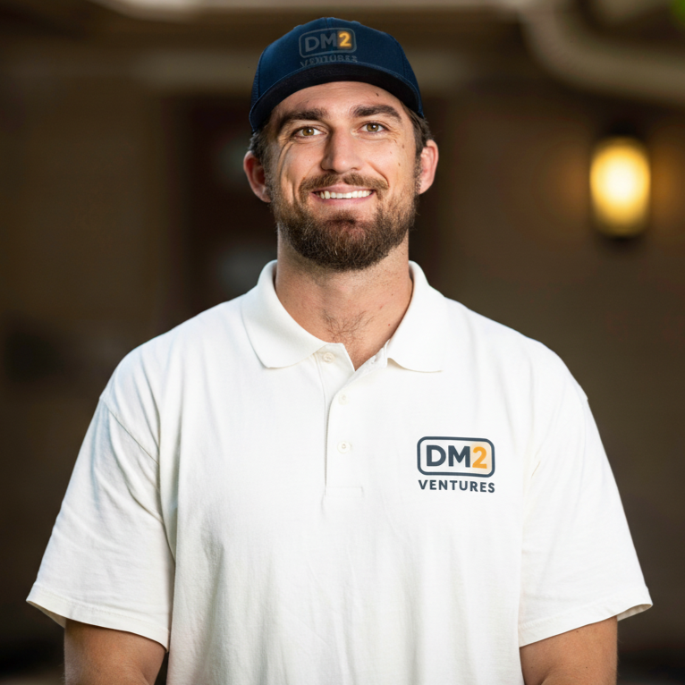 A man with a beard and a smile wearing a white polo shirt with a "DM2 Ventures" logo and a dark blue cap that also has a "DM2" logo.