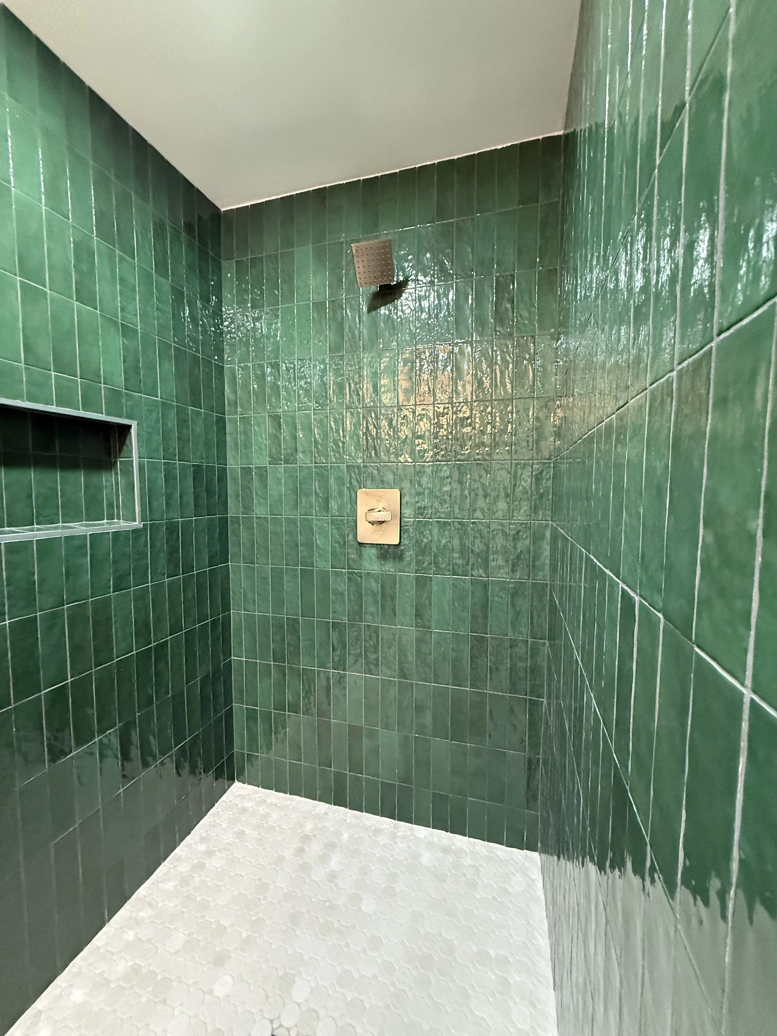 A shower with green tiled walls, a square showerhead, a built-in shelf, and a textured white tile floor.