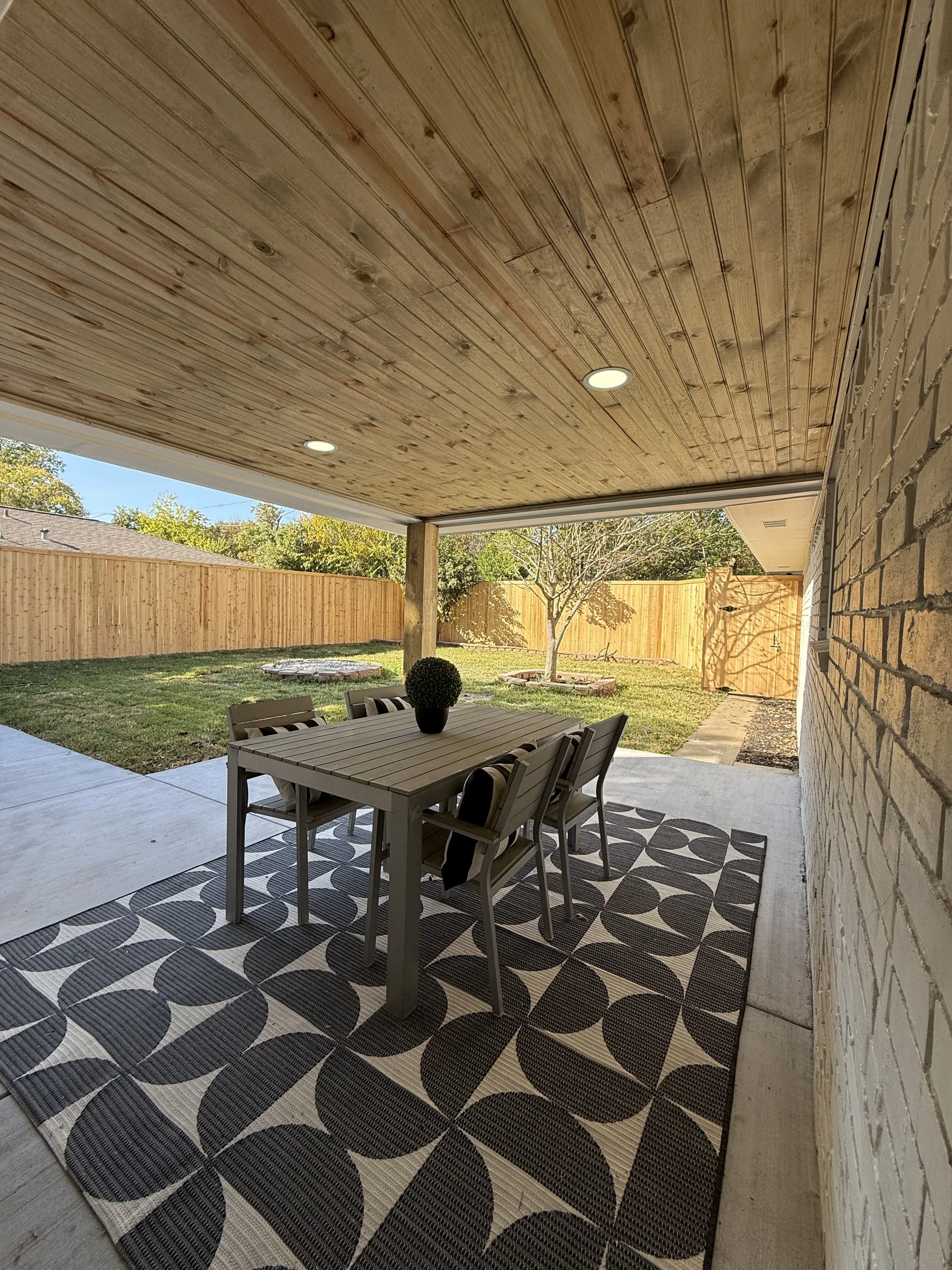 Covered outdoor patio with a dining table and six chairs, gray and black area rug, potted plant on the table, wooden ceiling, brick wall, backyard with grass, trees, and a wooden fence.