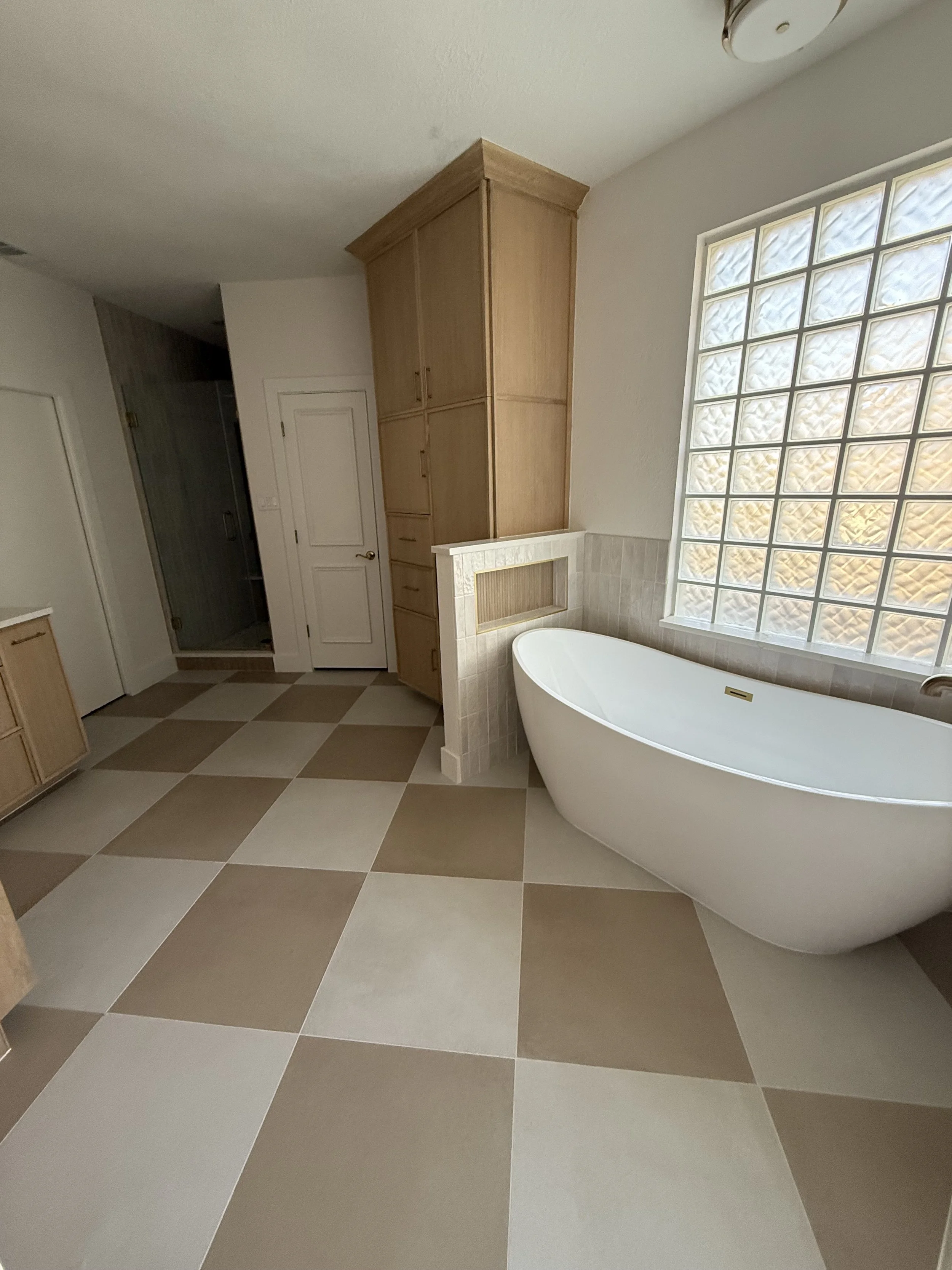 Bathroom with a freestanding bathtub near a large glass block window, and wooden cabinets with beige and brown checkered floor tiles.