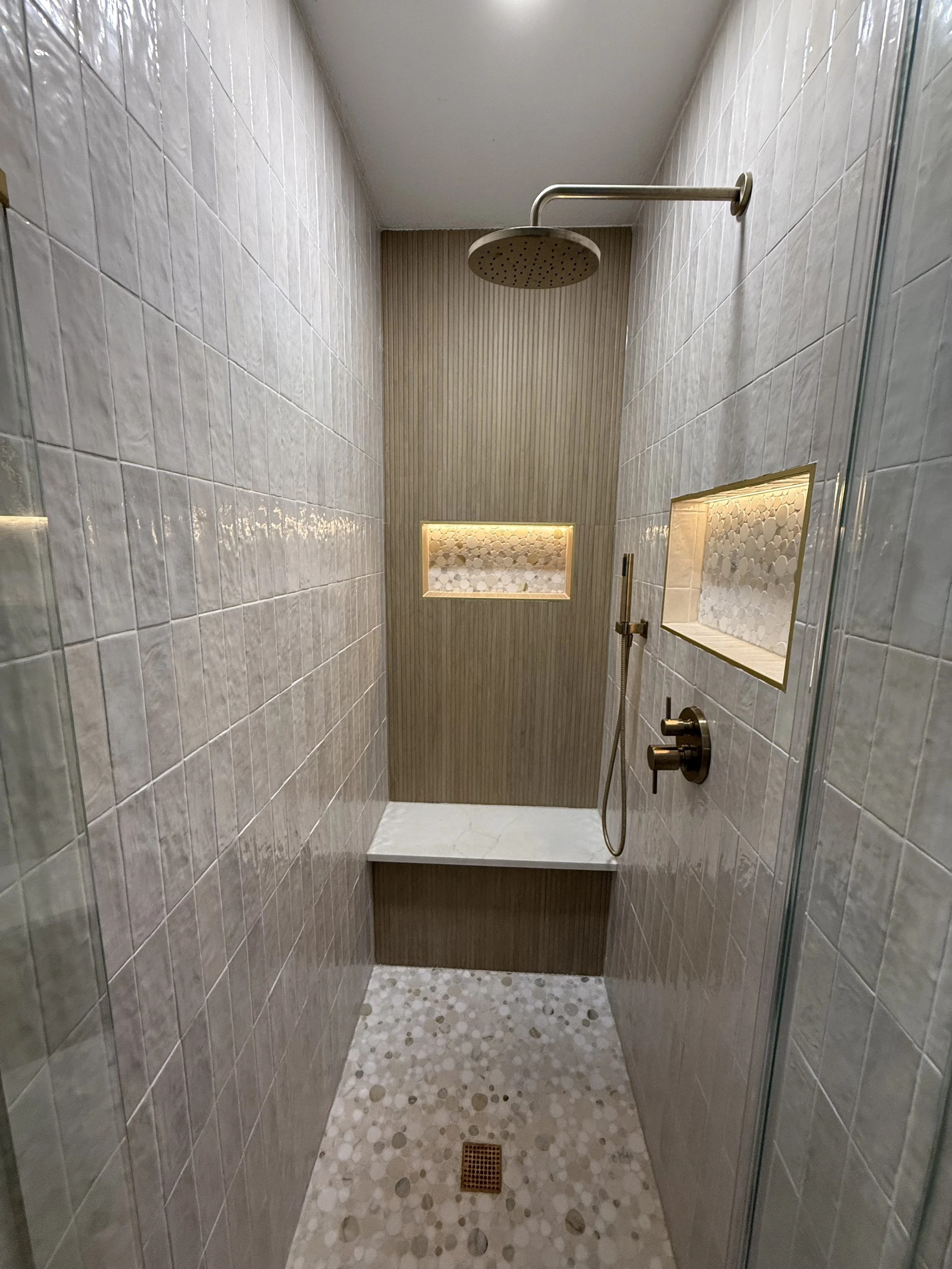 Shower with beige and gold tiled walls, built-in shelves, pebble floor, and a large overhead showerhead.