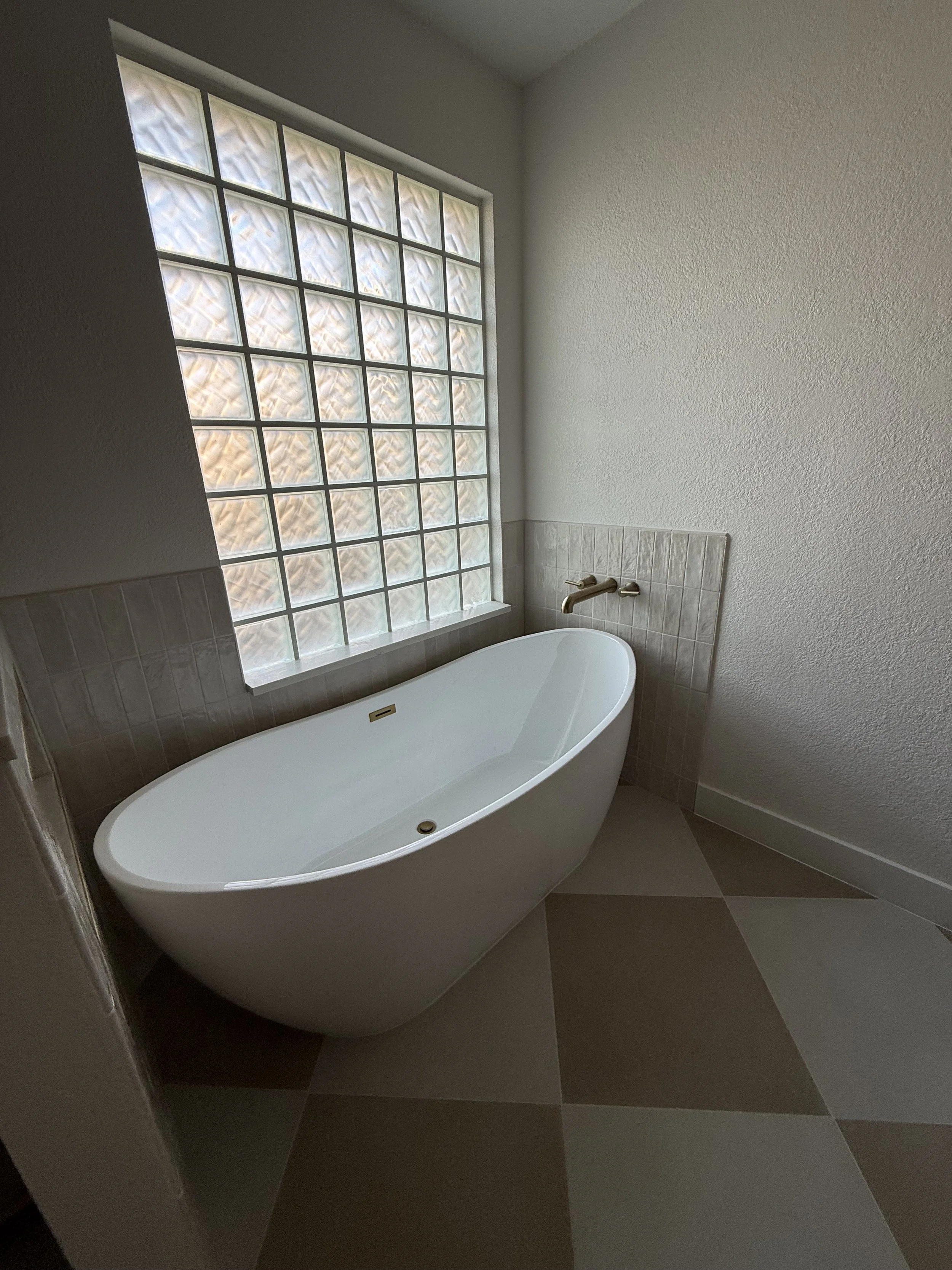 A modern bathroom with a standalone bathtub placed next to a glass block window for privacy and natural light, with beige tile flooring and a white textured wall.