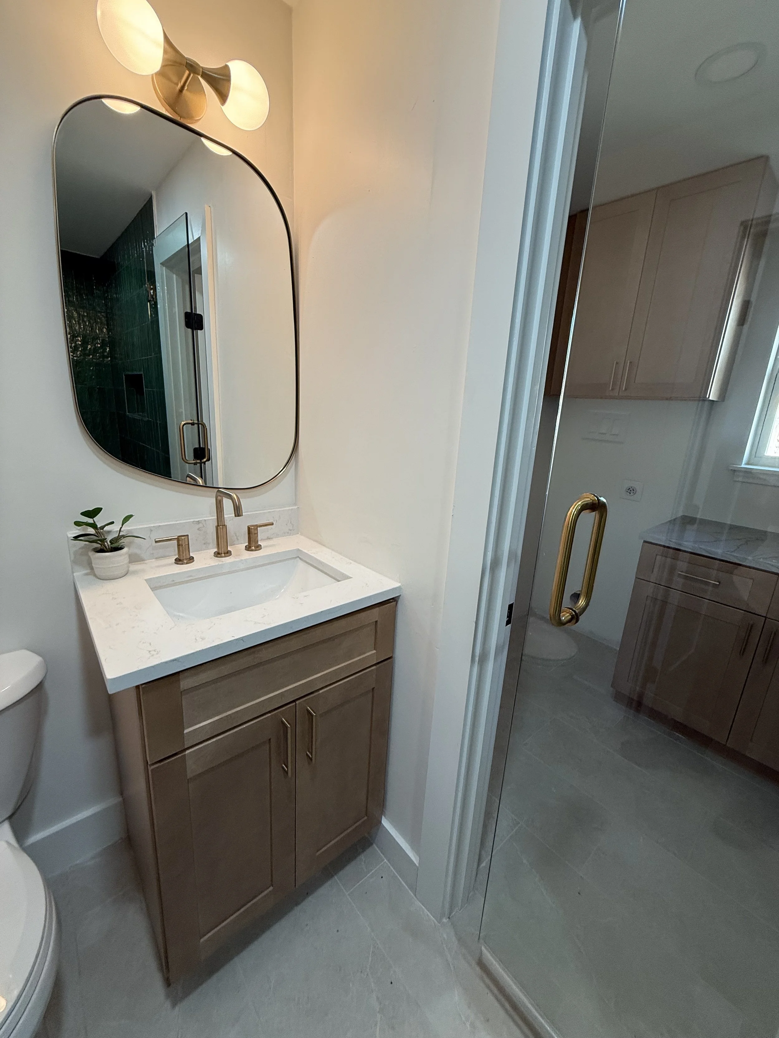 A small bathroom with a wooden vanity, white marble countertop, and a rectangular mirror with rounded edges. Above the mirror are two modern wall-mounted light fixtures. To the right, there is a glass shower door with a brass handle, and through the 