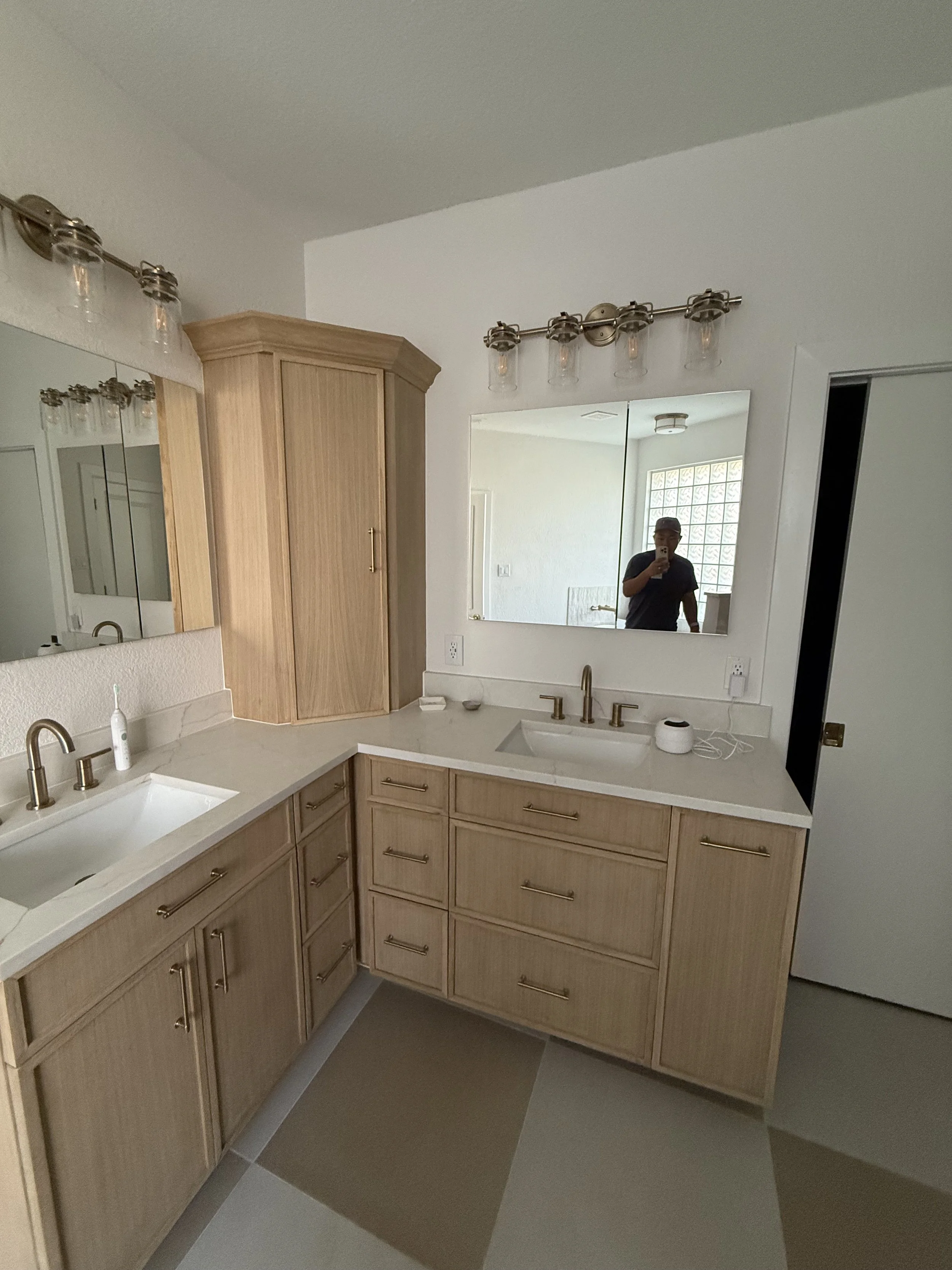 A bathroom with light wood cabinets, a white countertop, and two sinks with gold fixtures. A large mirror reflects a person taking a photo. The bathroom has a modern light fixture above the mirror and a window with glass blocks.