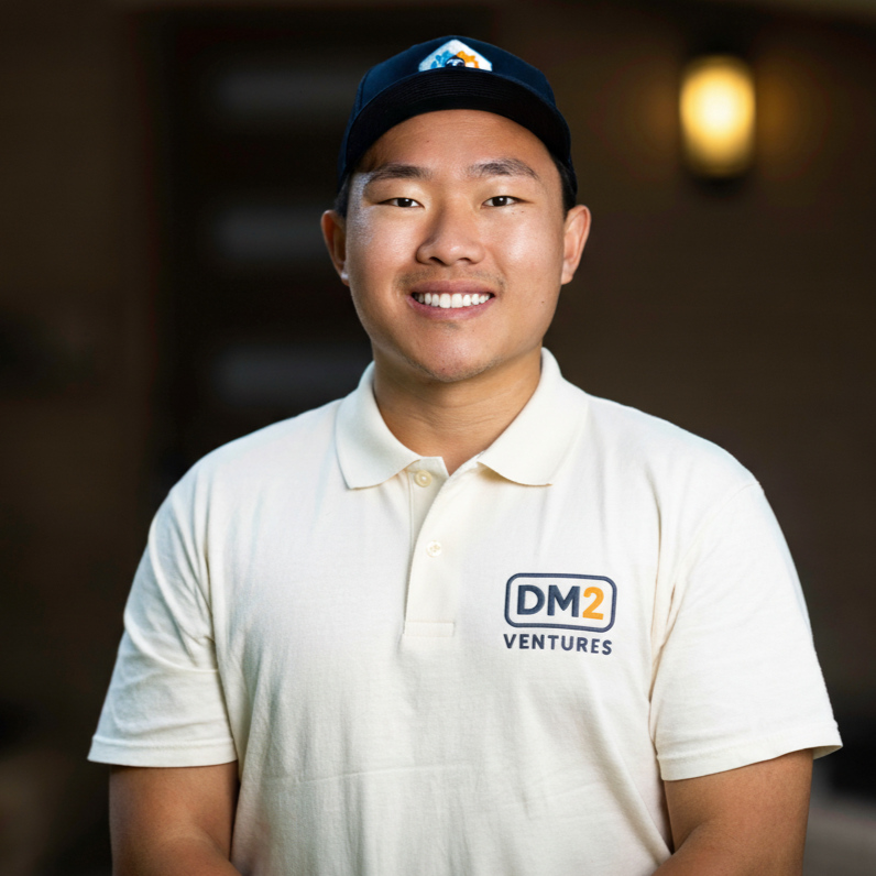 A young man smiling, wearing a white polo shirt with a logo that says 'DM2 Ventures' and a baseball cap with a mountain and sun logo, standing indoors with warm lighting.