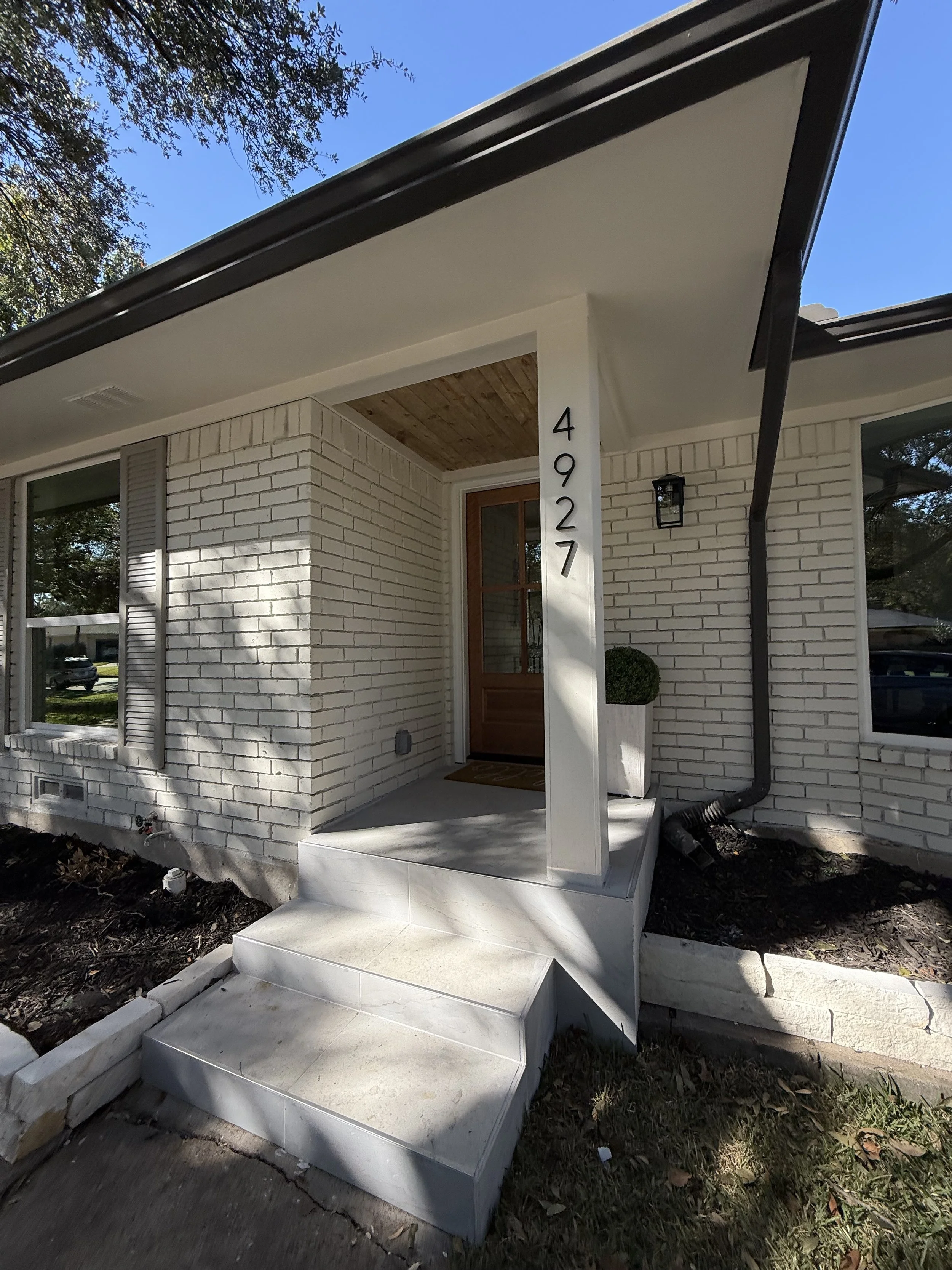 Front porch of a house with white brick walls, a wooden front door, and a set of three concrete steps leading to the entrance. The house number '4927' is vertically mounted on a white pillar. There are two windows, gutters, and a small planted shrub 