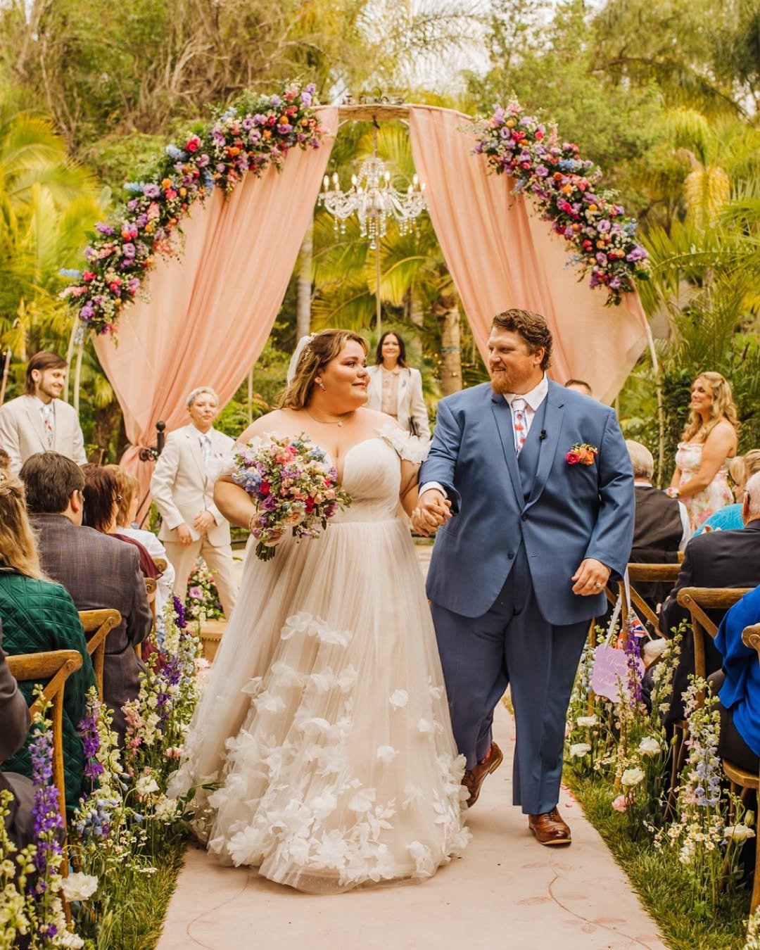 Couple walking down the aisle after their wedding with floral arch in background and wedding guests applauding