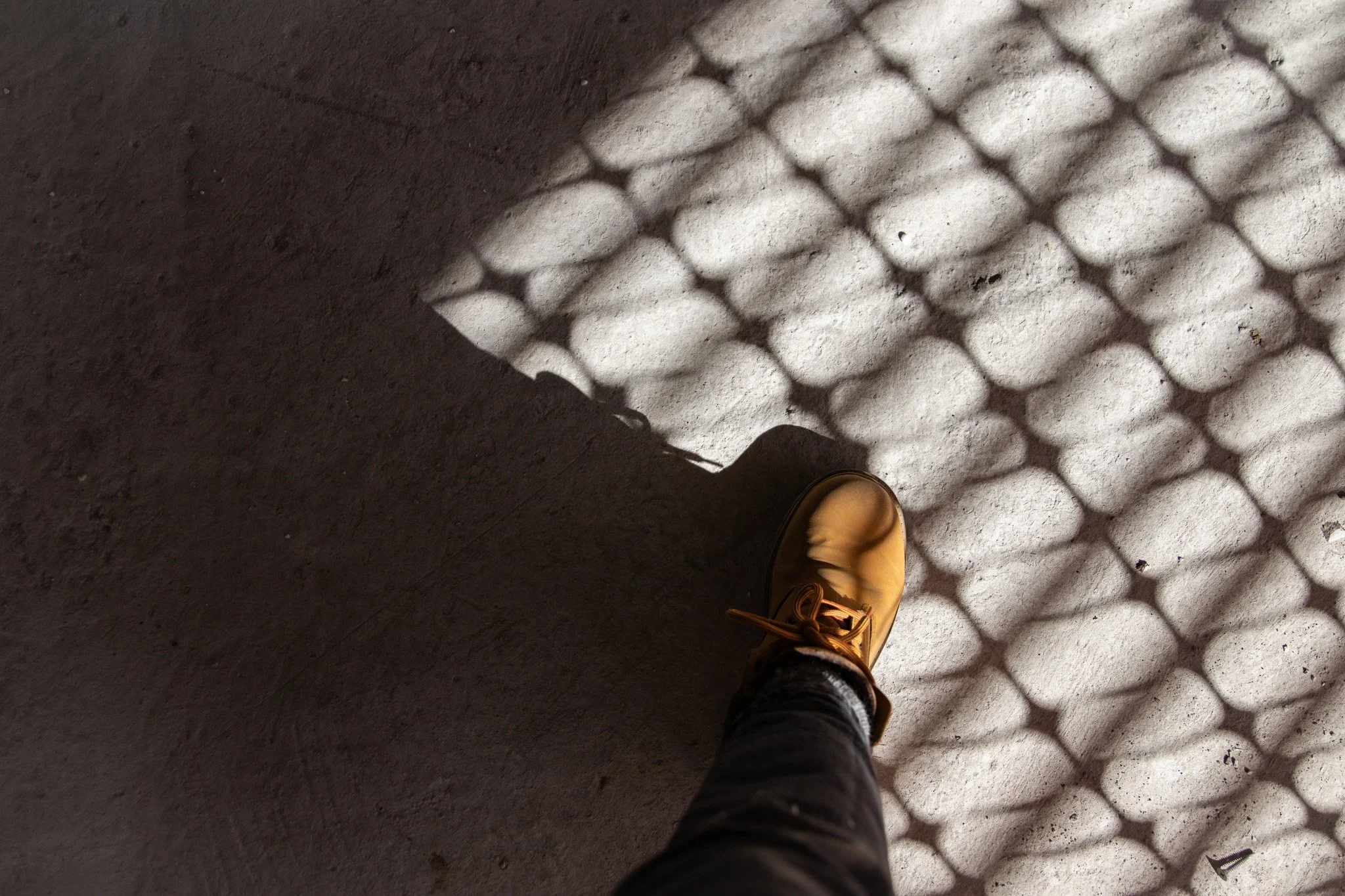 Una sombra de reja en el suelo de concreto, con el pie de una persona usando un zapato color camel y pantalones oscuros.