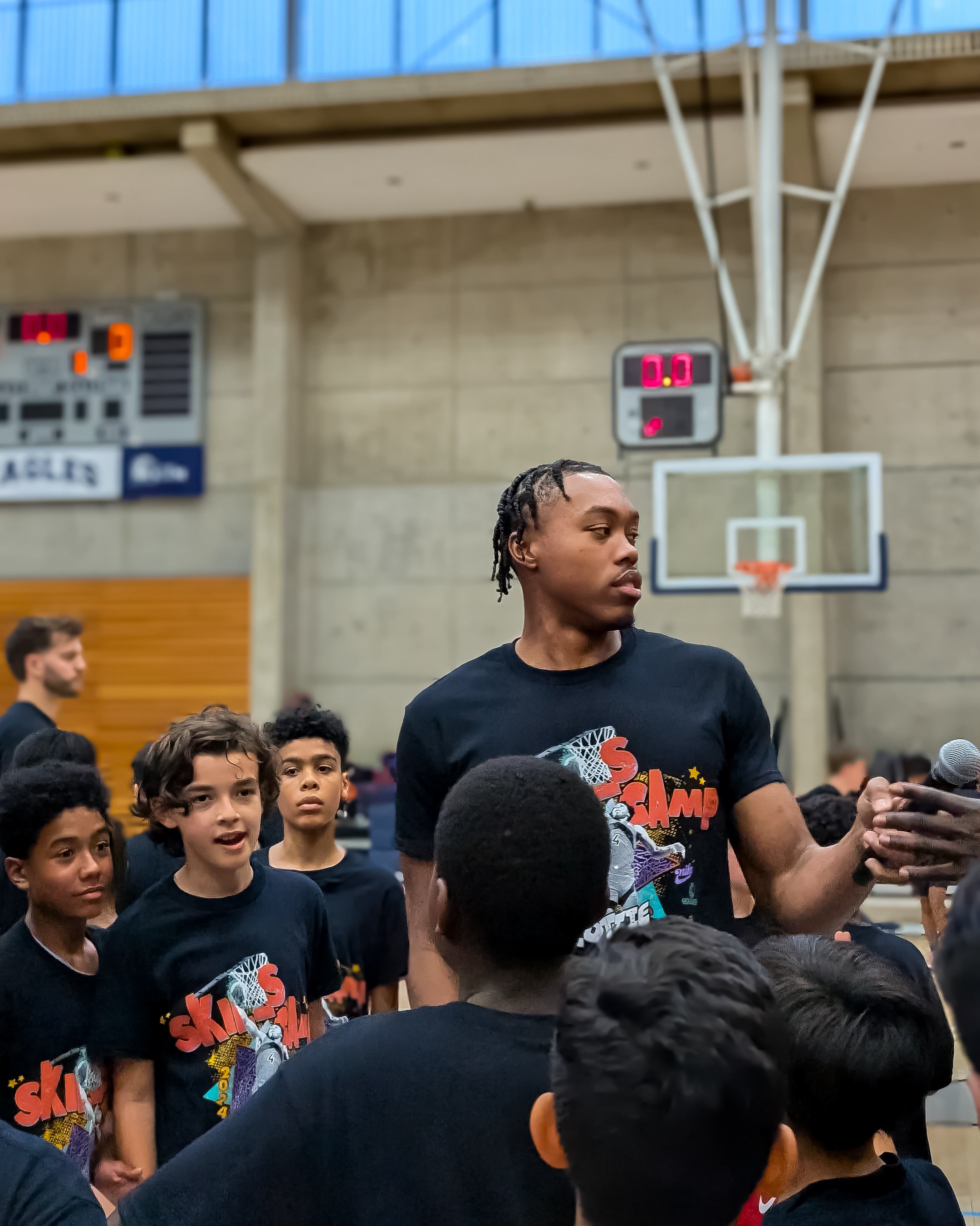 A man surrounded by a group of young people in a gymnasium, all wearing matching T-shirts with a graphic design. There's a basketball hoop and scoreboard in the background. The man appears to be speaking or addressing the group.