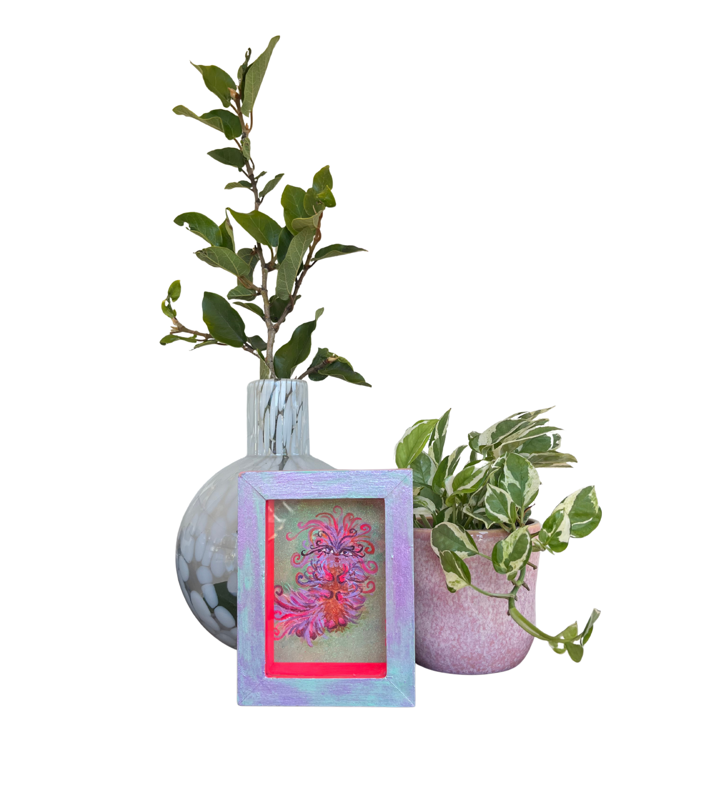A still life arrangement of two potted plants, a framed artwork, and a glass vase with green foliage. The taller plant has dark green leaves, and the shorter plant has variegated green and white leaves. The framed artwork in front features a colorful