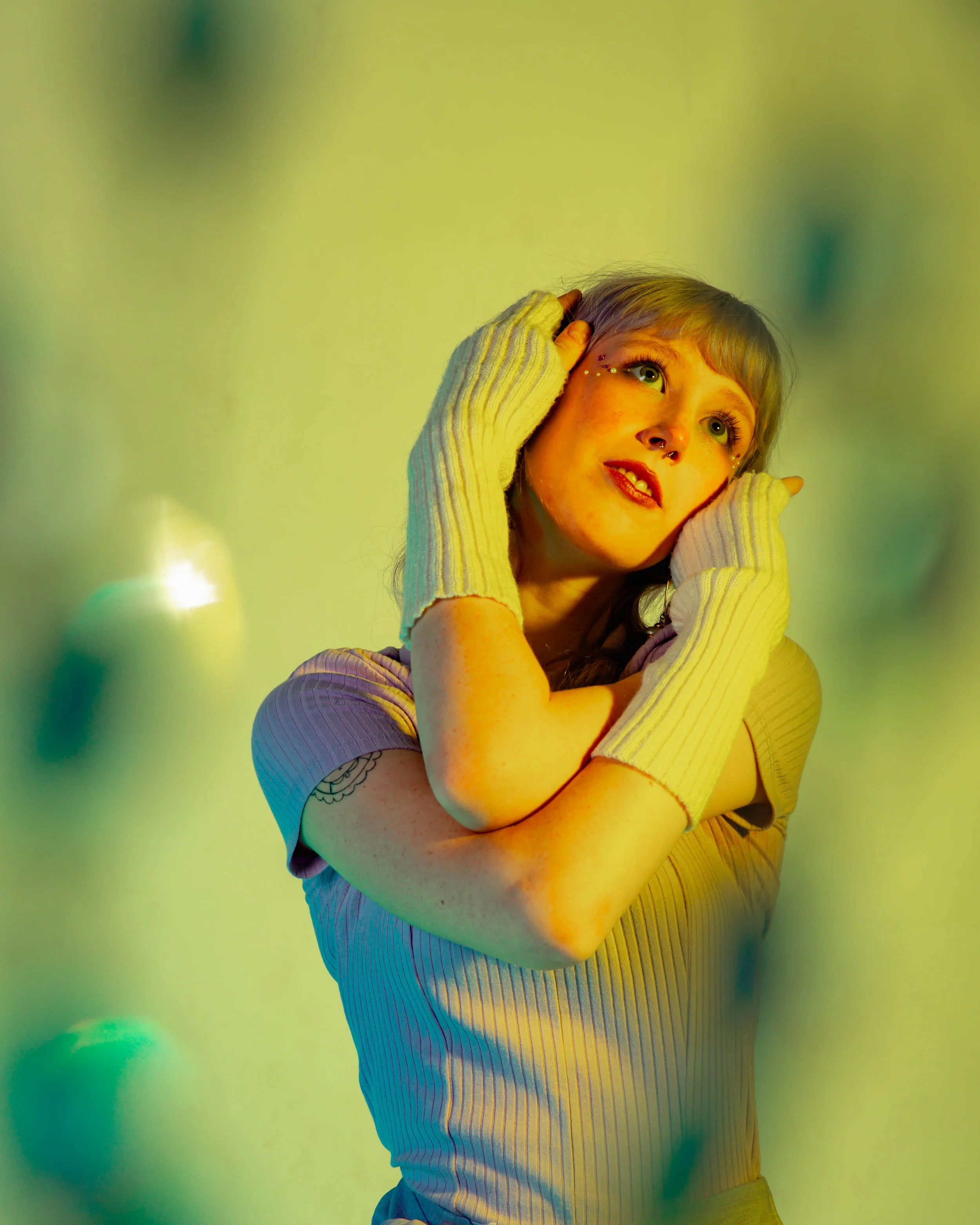 A young woman with short blonde hair, green eyes, and freckles, wearing a ribbed lavender shirt, yellow arm warmers, and glitter makeup, posing against a blurred green background with bokeh effects, touching her head and looking upwards.