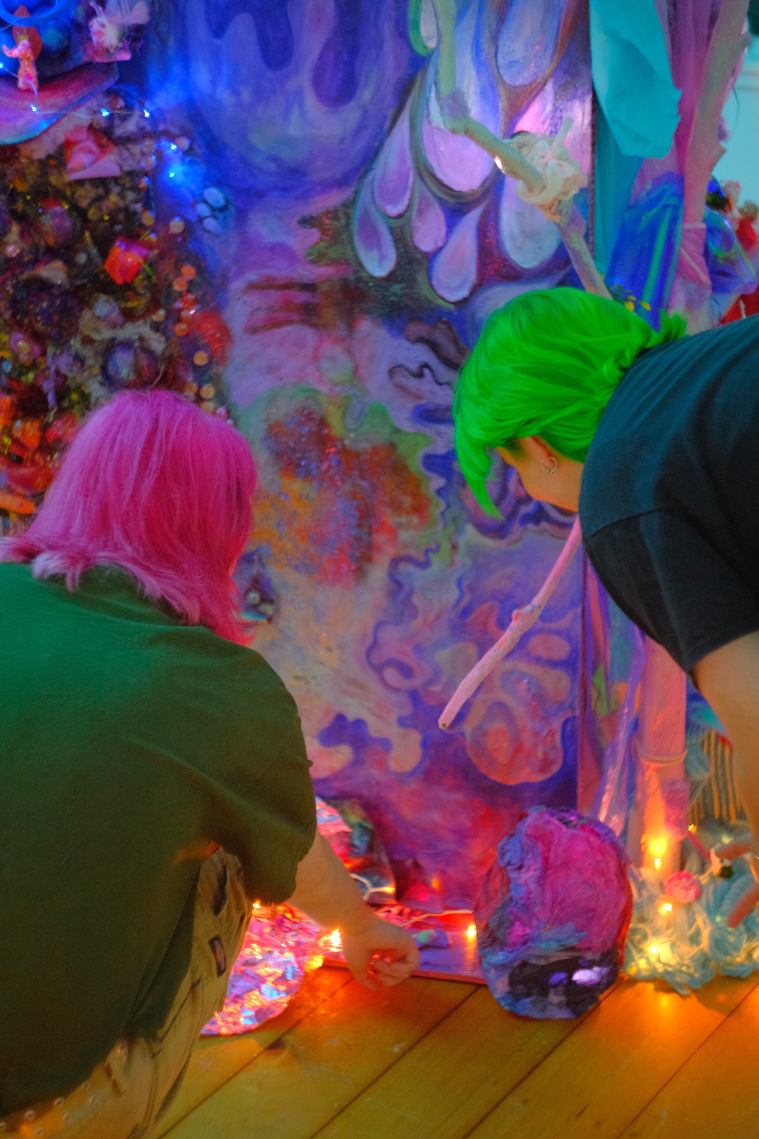 Two people with brightly colored hair, pink and green, are decorating a vibrant, abstract, and swirling mural with various objects and lights on the floor.