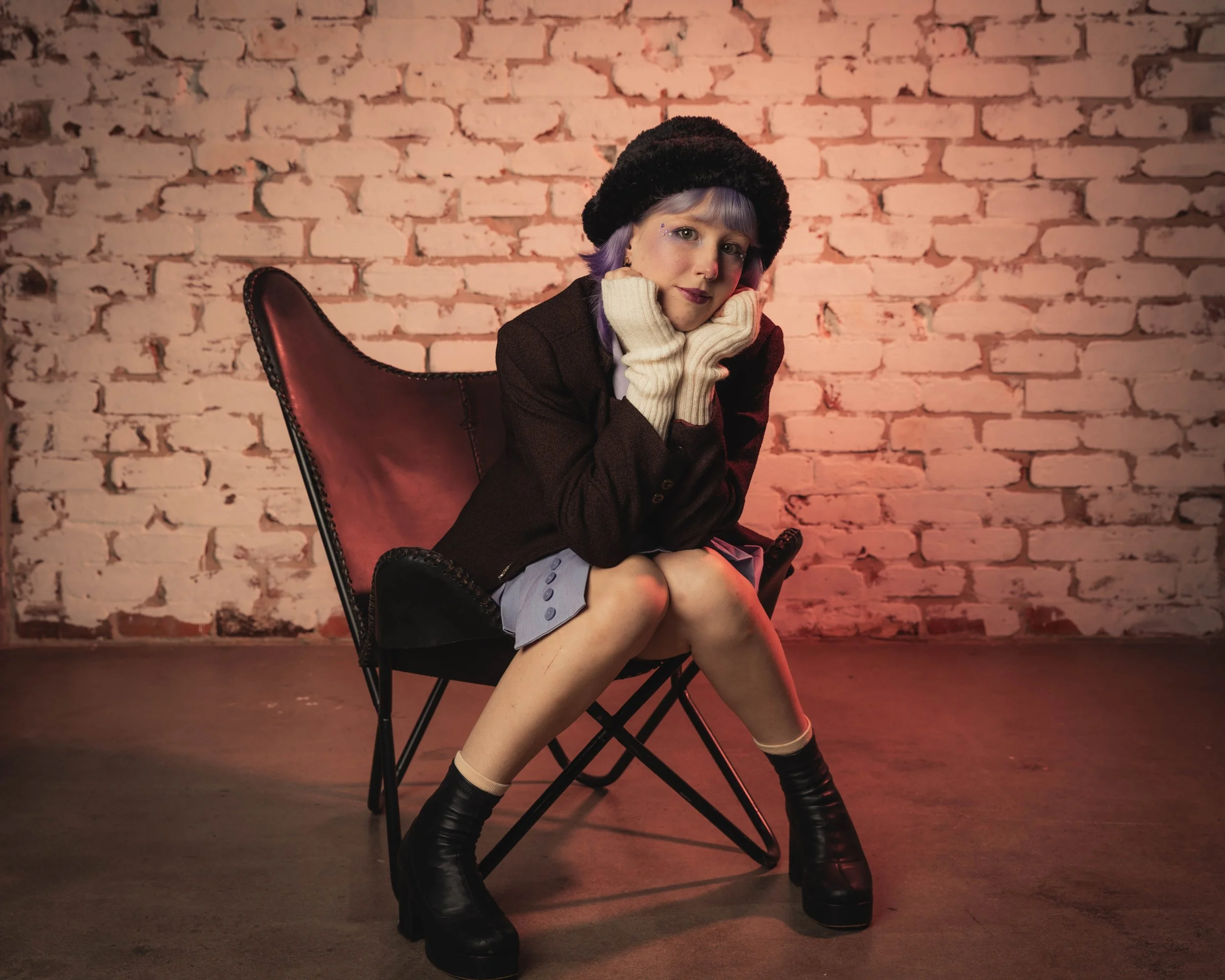 A young woman with purple hair sitting on a leather chair against a brick wall, wearing a black beret, brown blazer, white gloves, gray skirt, and black boots.
