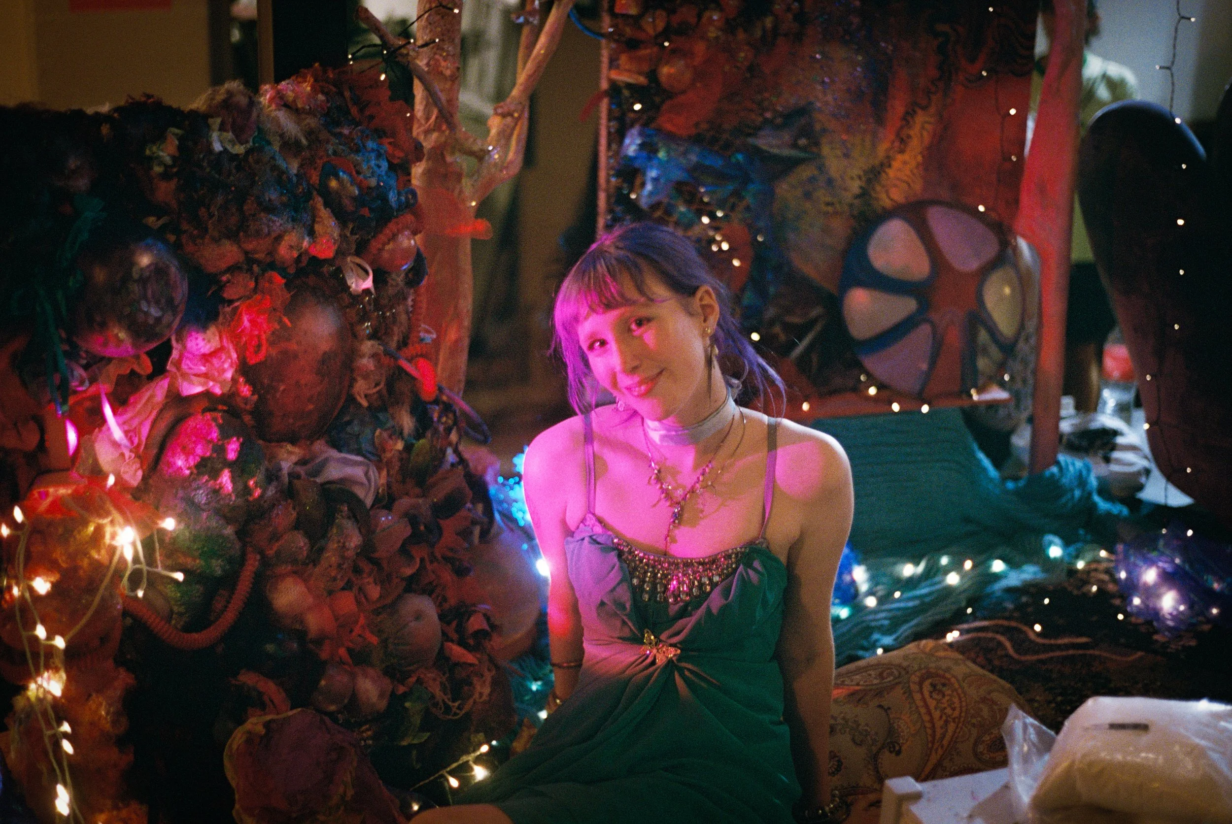 A young woman with purple hair and a choker necklace, smiling and sitting on the floor at a decorated party or celebration with colorful lights, art pieces, and decorations around her.
