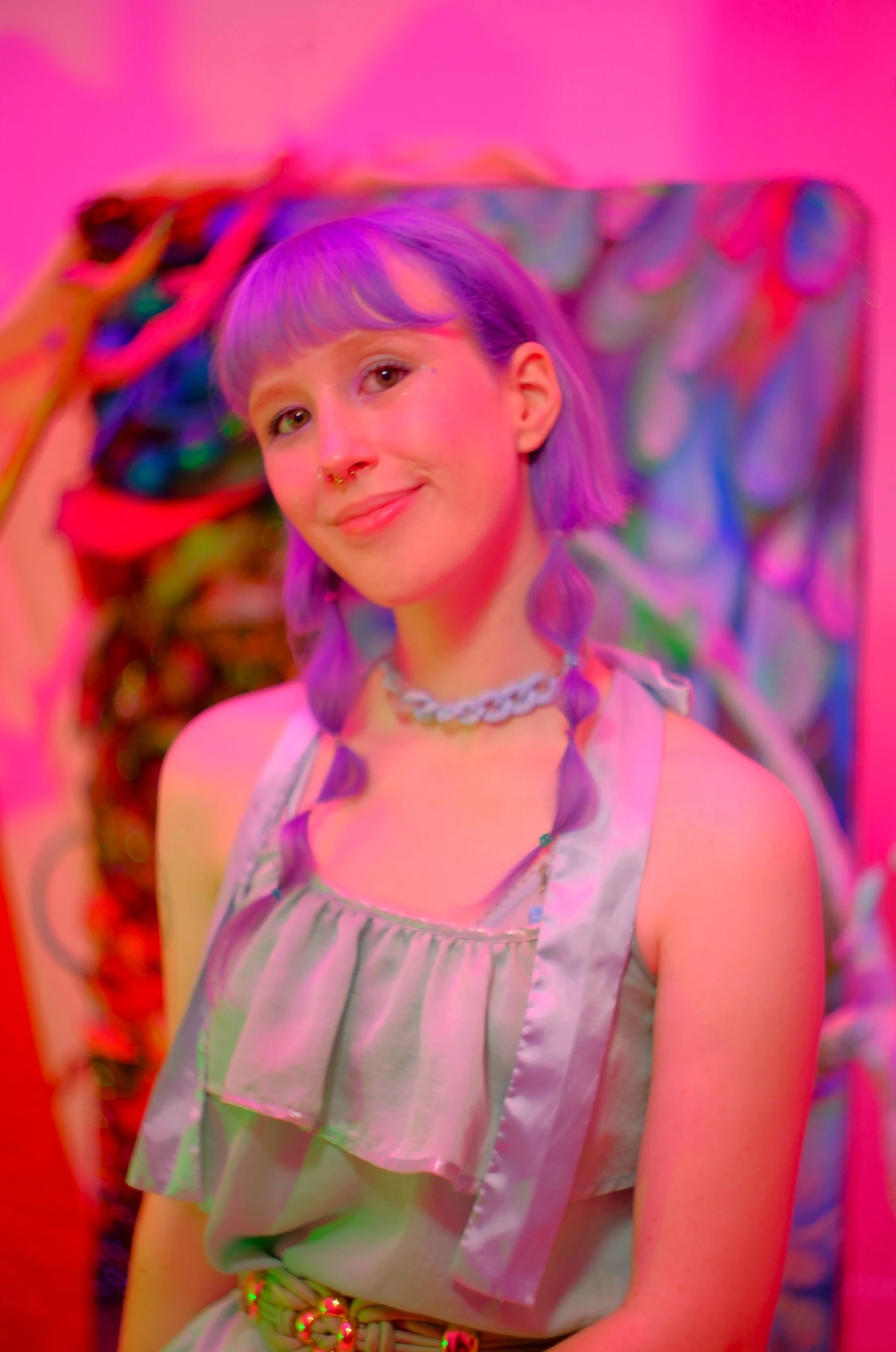 Young woman with purple hair and septum piercing, wearing a gray sleeveless top and chain necklace, sitting against colorful abstract art with pink lighting.