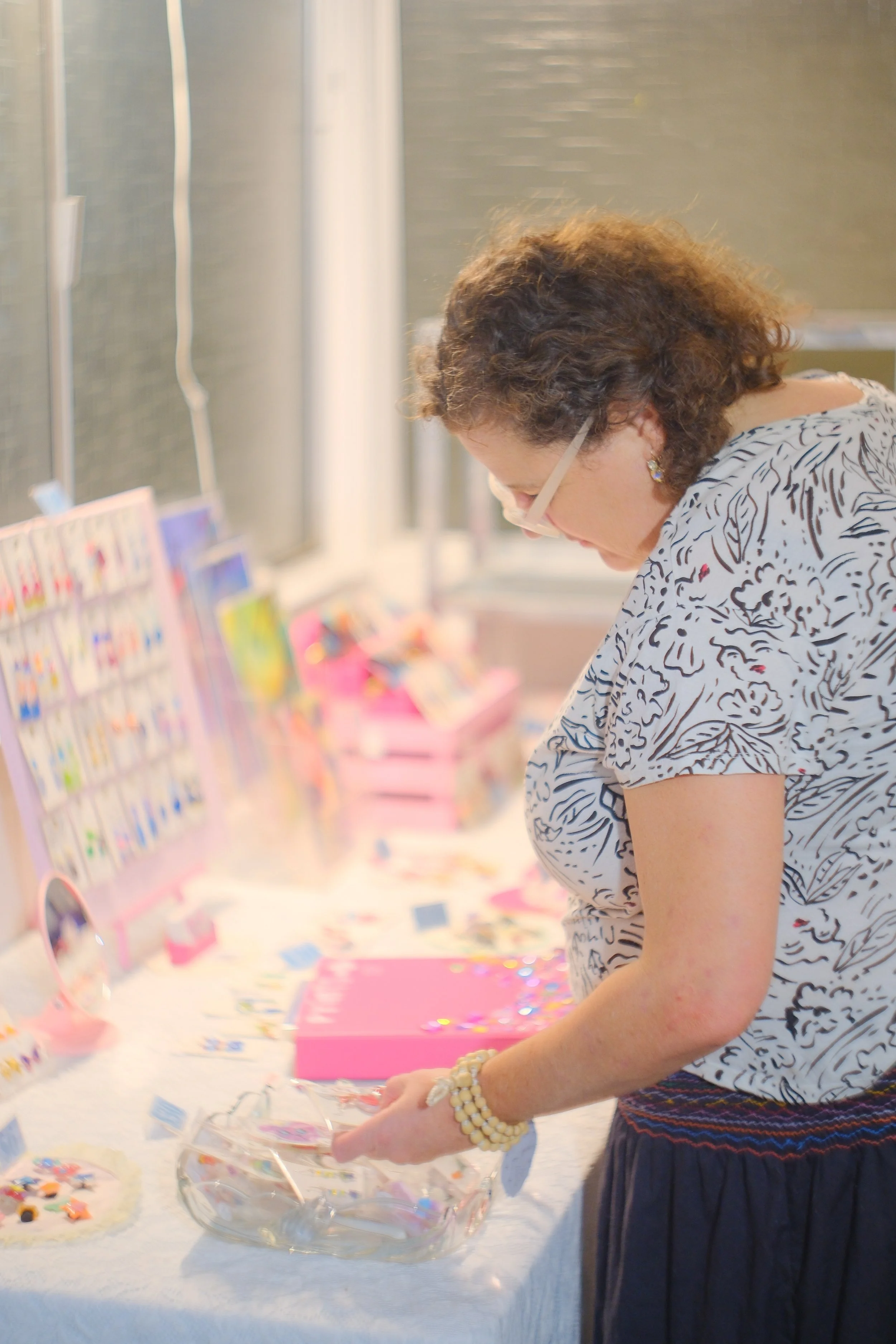 Woman with curly hair in glasses, wearing a printed white top and dark skirt, browsing colorful accessories and jewelry display at a craft fair or boutique.