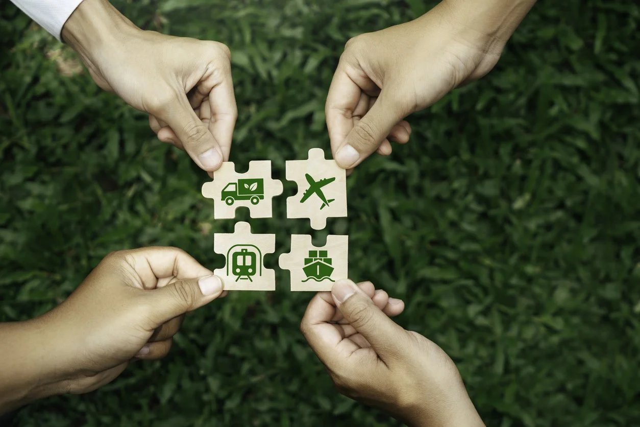Four hands holding puzzle pieces with eco-friendly transportation and travel icons over a grassy background.