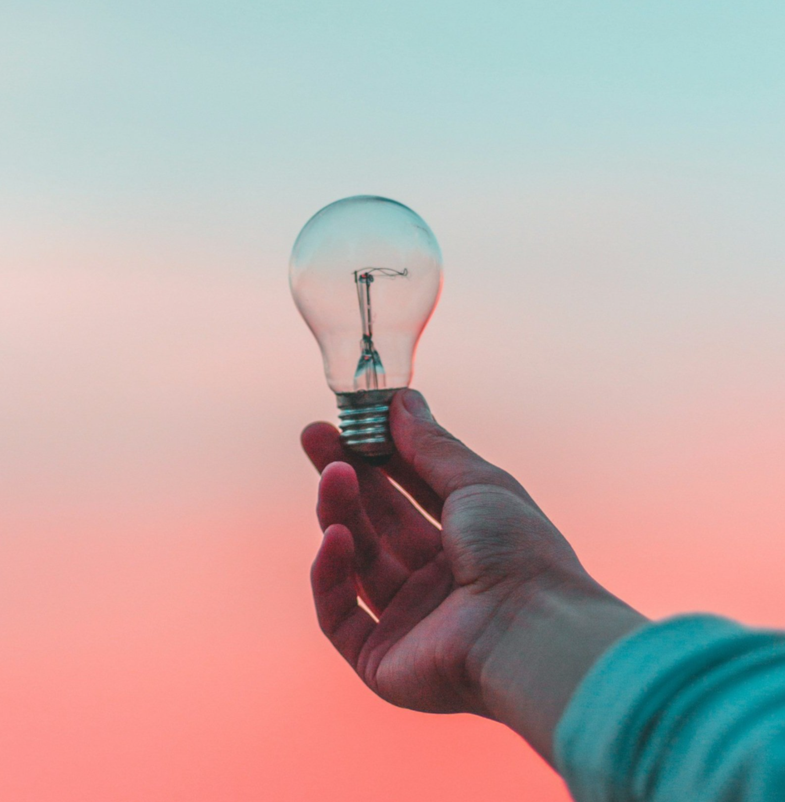 A person's hand holding a clear light bulb against a pink and blue gradient sky.