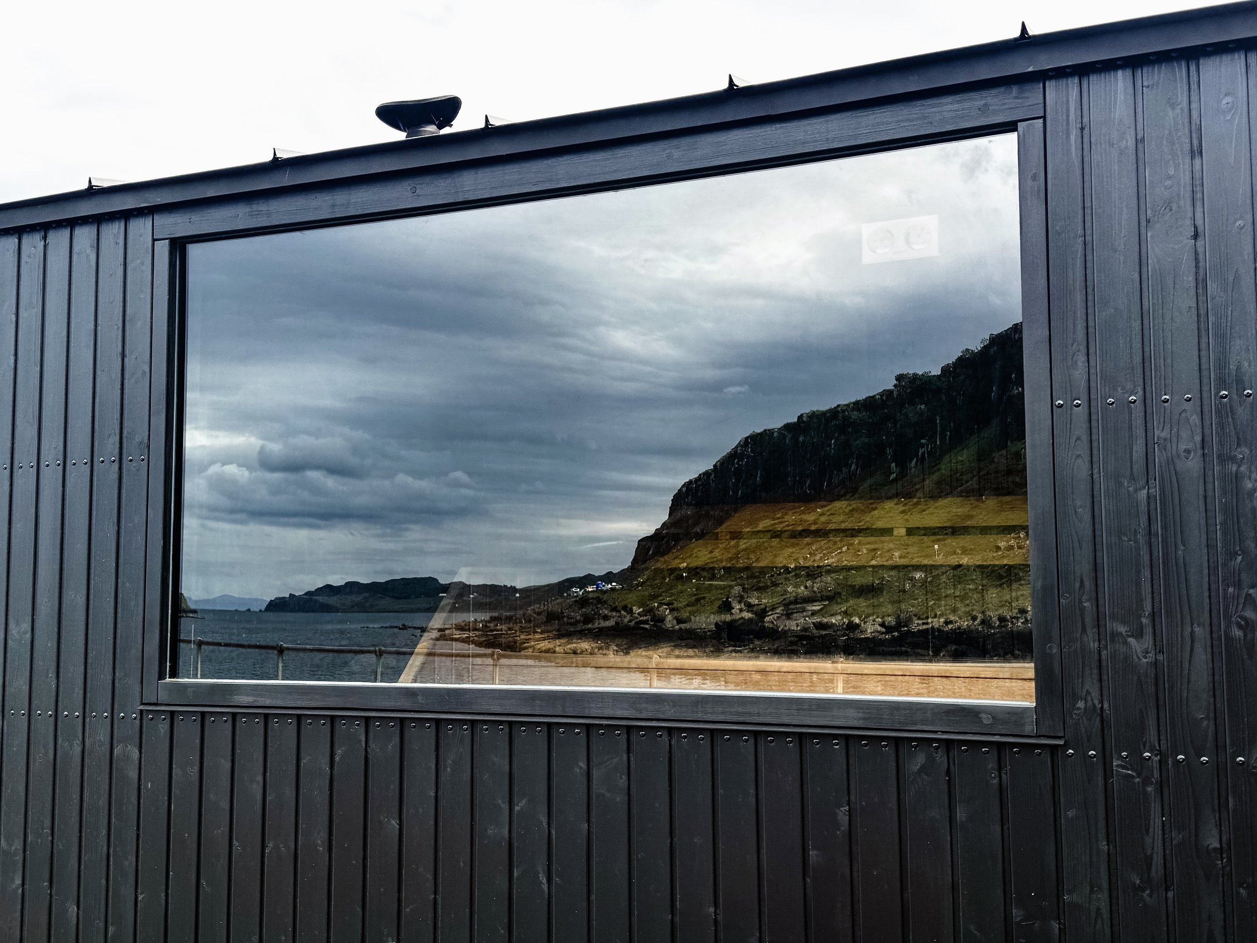 Interior of a wooden sauna with a wood-burning stove, benches, and a large window. Warm up after a wild swim and escape from the ever-changing weather on the Isle of Skye at Staffin Sea Sauna on Isle of Skye 