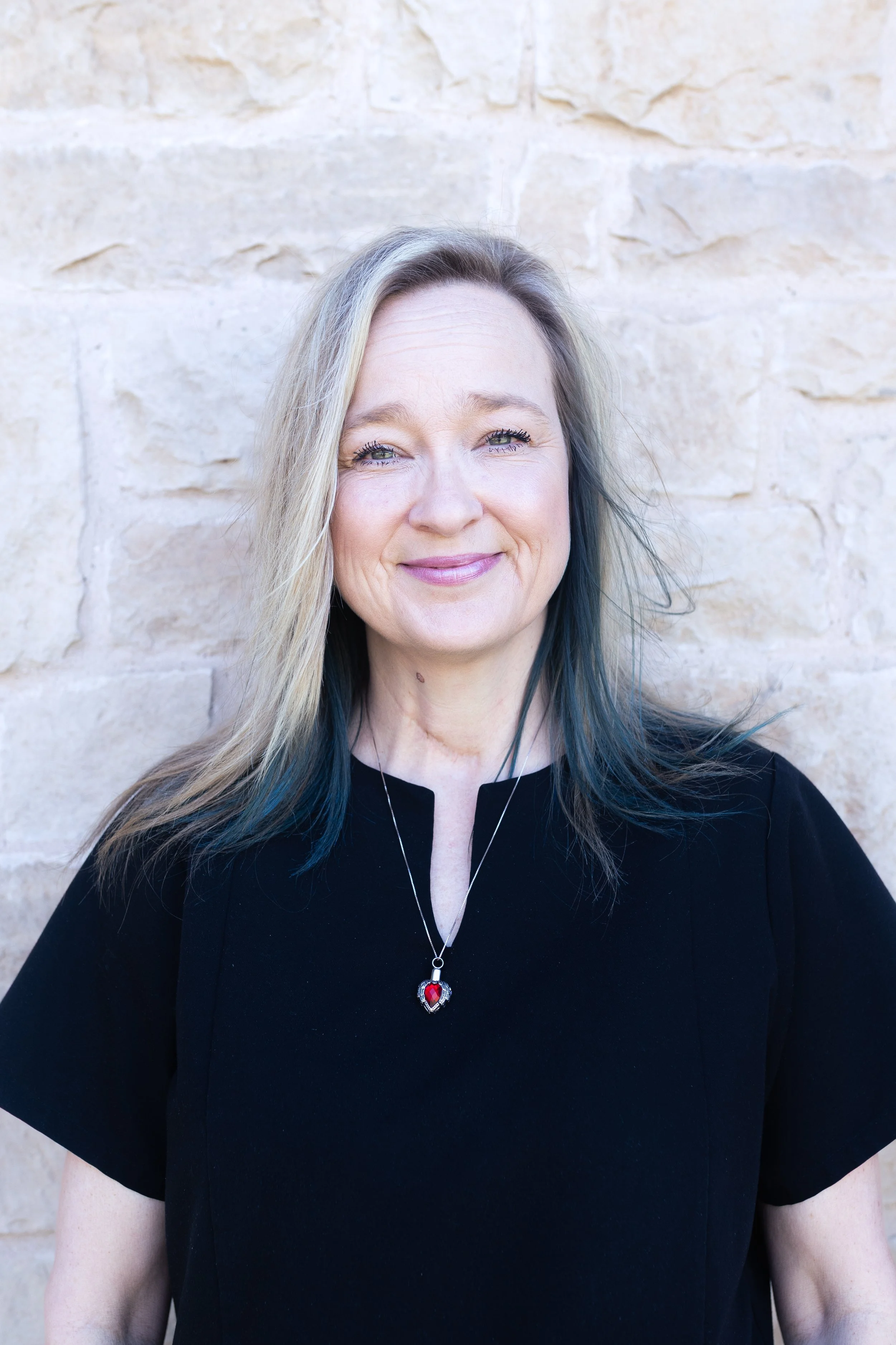 Portrait of a smiling woman with blonde and light blue hair, wearing a black top and hoop earrings, against a neutral background.
