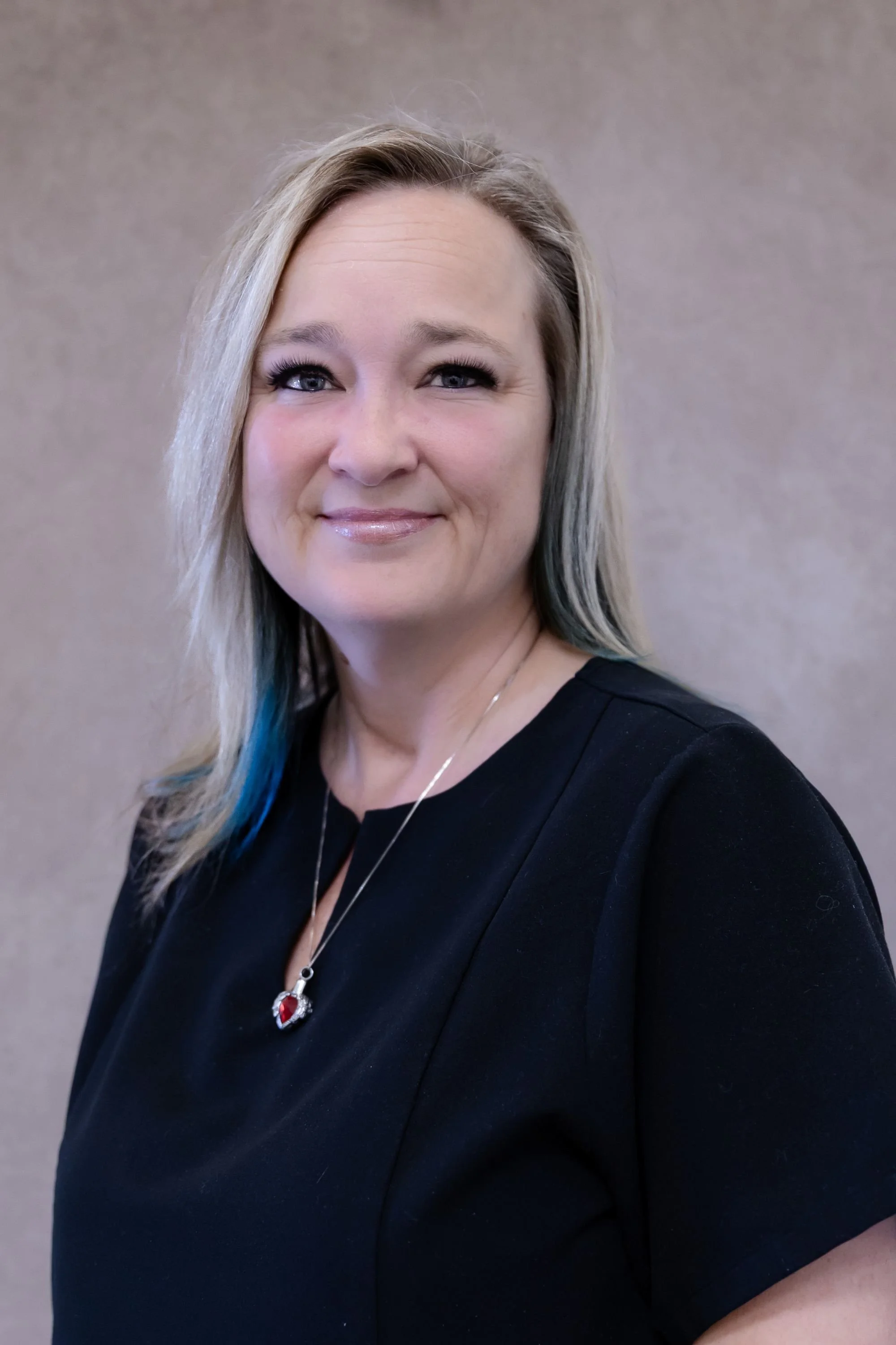 Portrait of a smiling woman with blonde and light blue hair, wearing a black top and hoop earrings, against a neutral background.