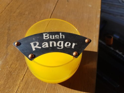 Small glass with "Bush Ranger" moustache protector, on wooden surface.