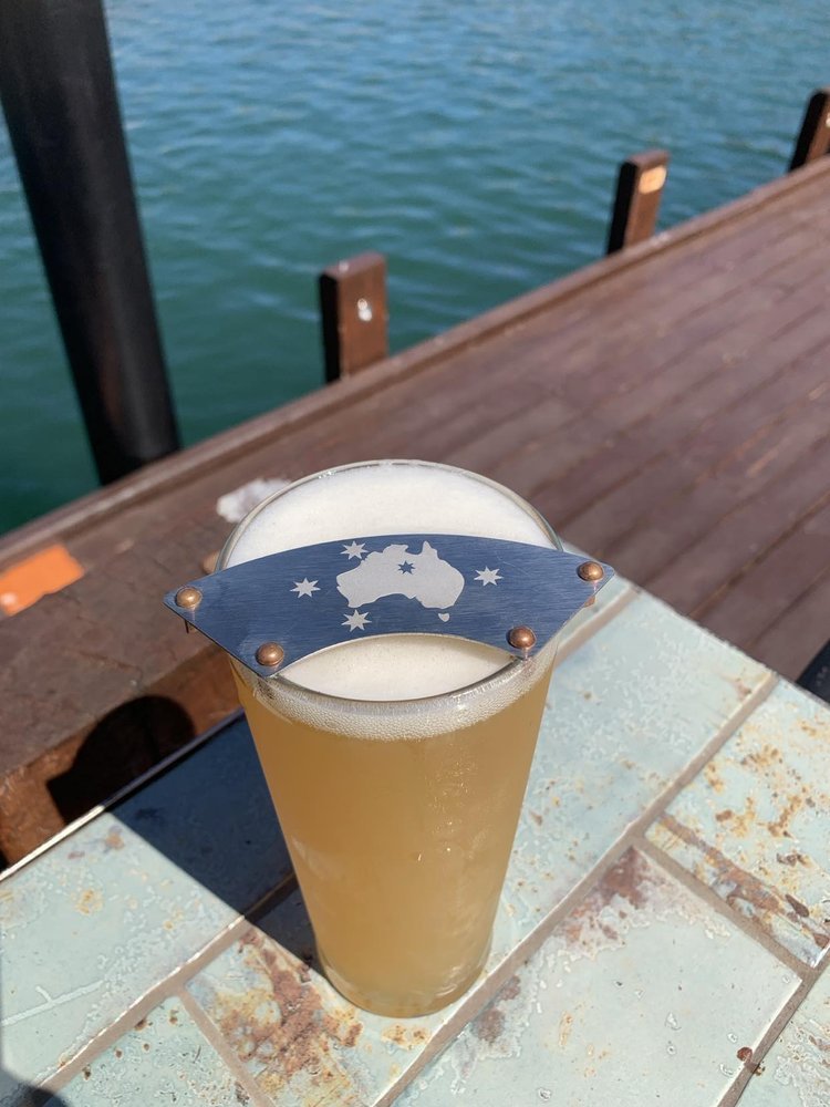 Glass of beer with a metal moustache protector featuring Australia and southern cross, placed on a table by the water.