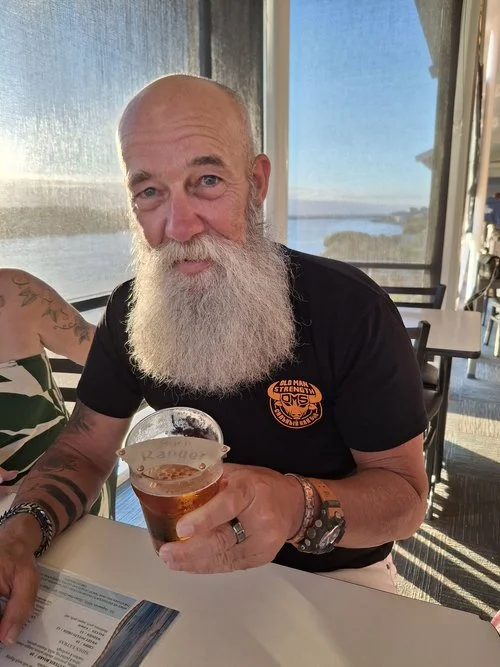 A bearded man holding a drink at a sunny indoor dining area.