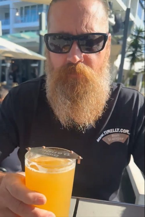 Bearded man wearing sunglasses holding a glass of beer at an outdoor cafe.
