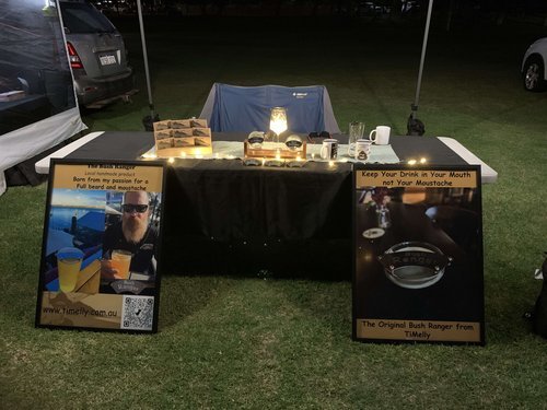 Outdoor night with two signs advertising beard-related product from "Melly." The signs feature images of a beard product designed to prevent drinks from spilling into moustaches. The booth is illuminated with small lights.