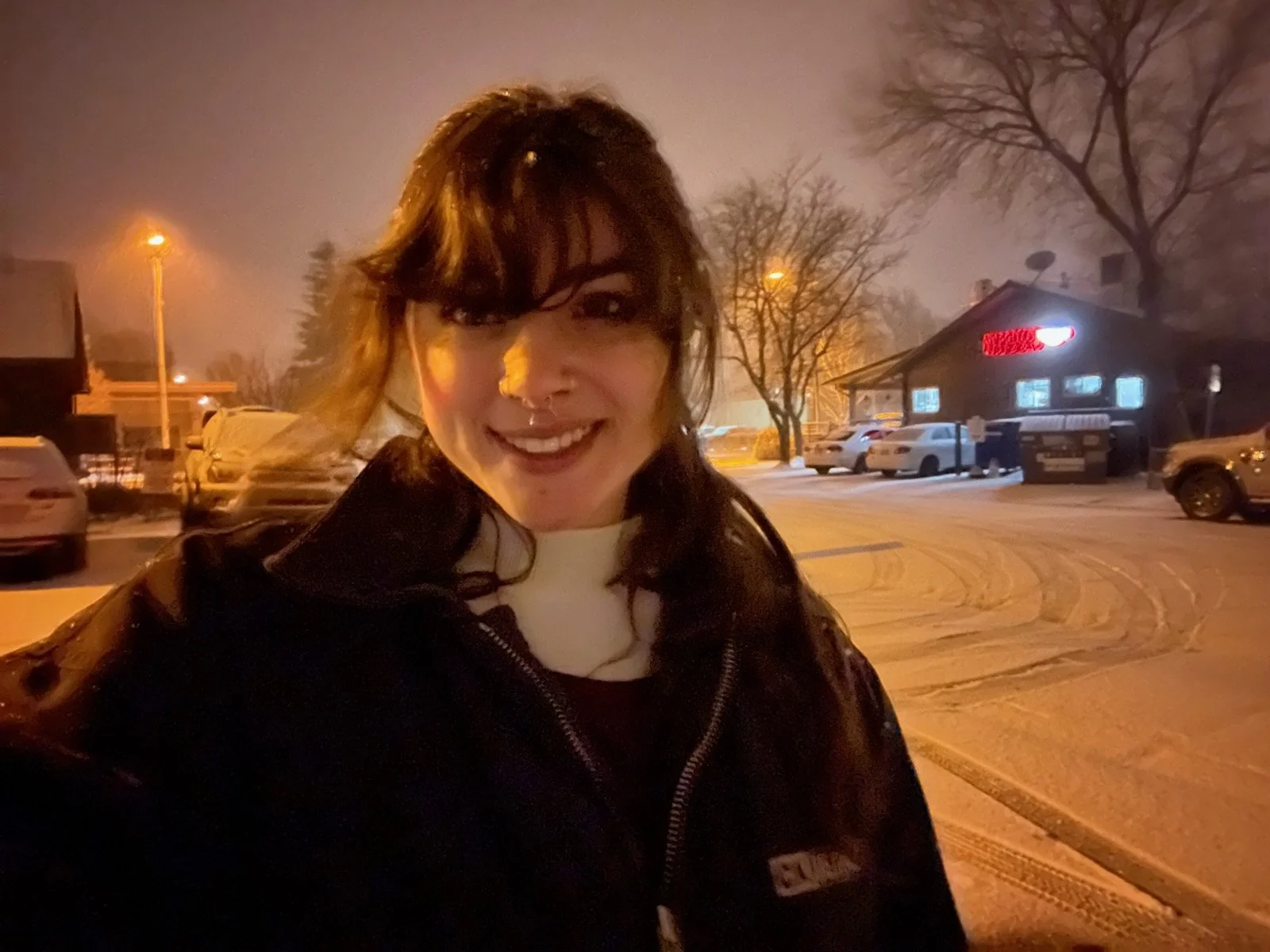 A woman with dark hair and a nose ring smiling outdoors at night in a snowy parking lot with trees, cars, and a building with a red sign in the background.