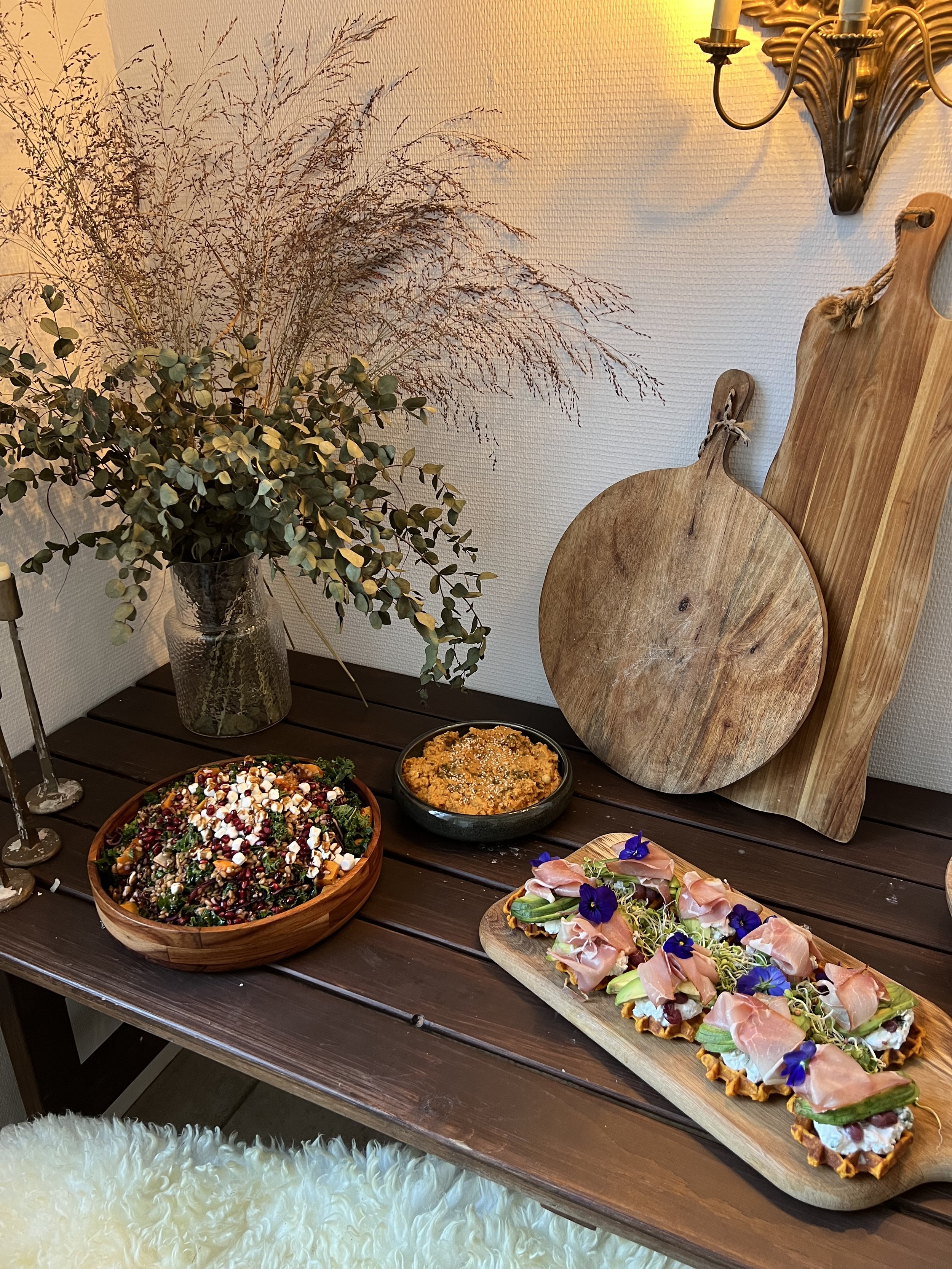 A wooden table with a rustic food spread, featuring a salad in a wooden bowl, a dish of couscous, and a platter of appetizers with prosciutto and edible flowers. Decorative grass in a vase and wooden cutting boards are in the background, along with a wall-mounted candle holder.