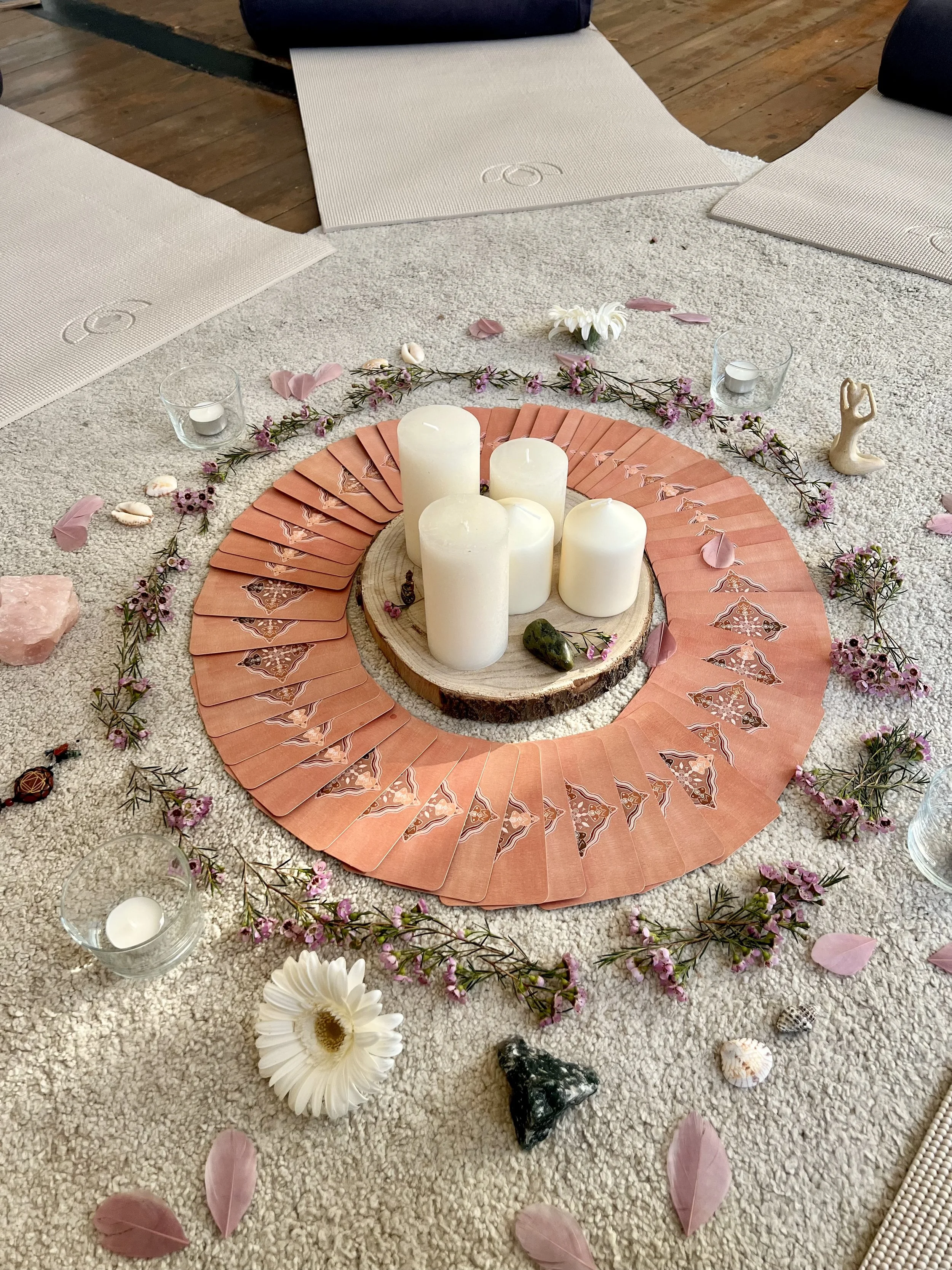 A women's circle setup featuring five white candles on a wooden disc surrounded by a circle of tarot cards, flowers, and small objects like crystals and shells, placed on a light carpet. Yoga mats are positioned around the arrangement.