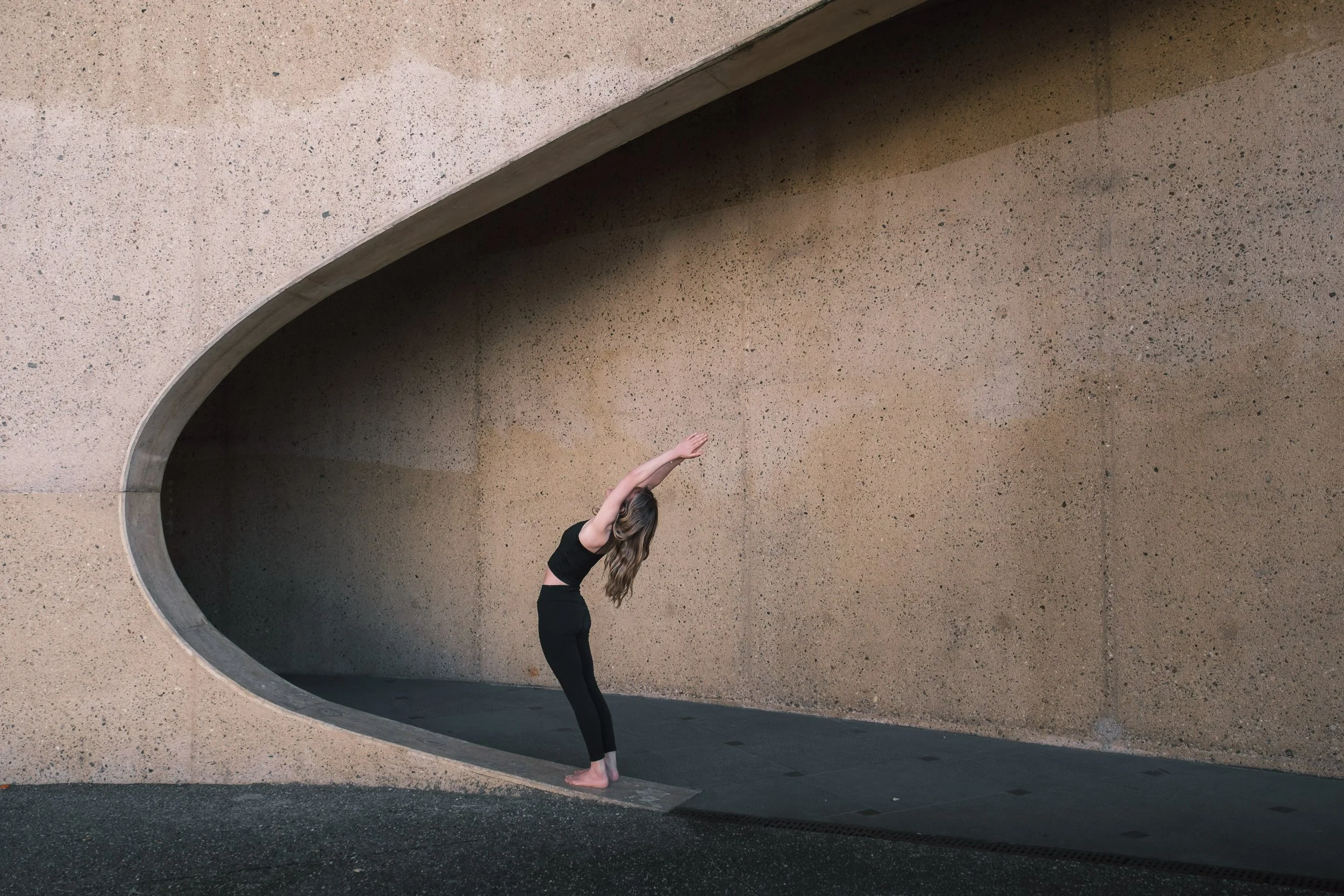 Person practicing yoga in athletic attire, performing a backbend under a concrete architectural structure.