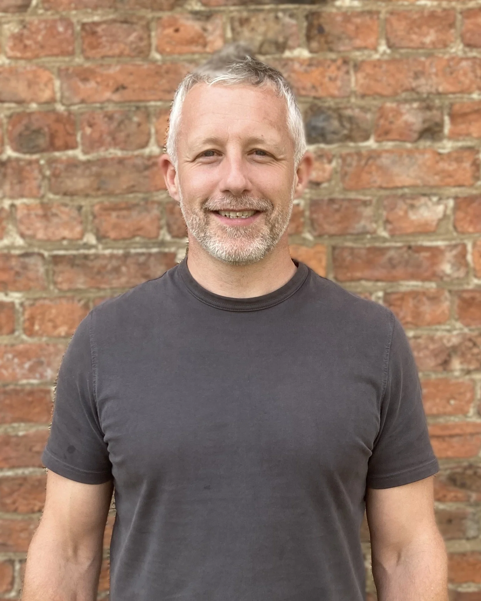 man with a beard in dark grey t shirt in front of a red brick wall