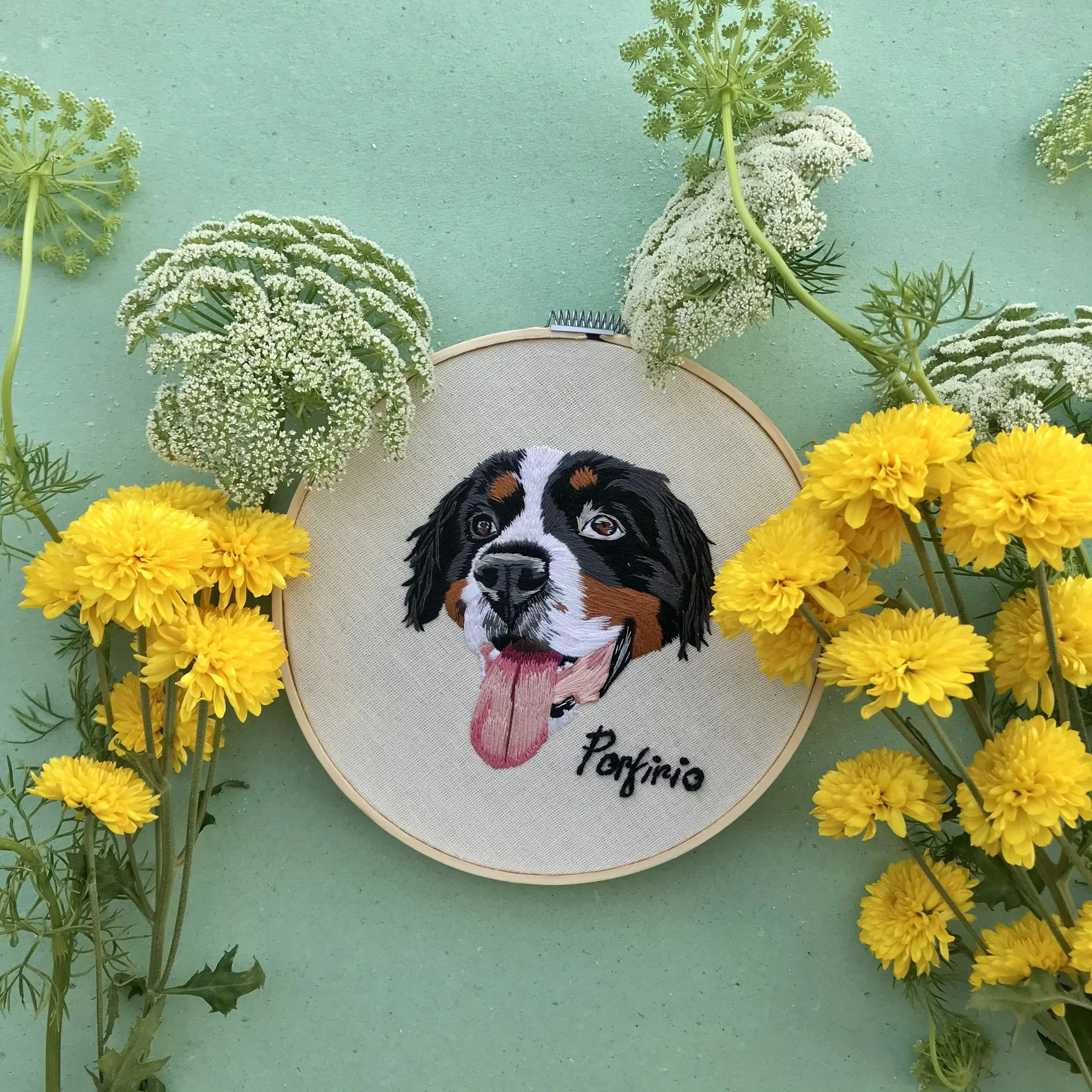 Un ricamo di un cane con la lingua fuori, intorno a fiori gialli e bianchi su sfondo verde