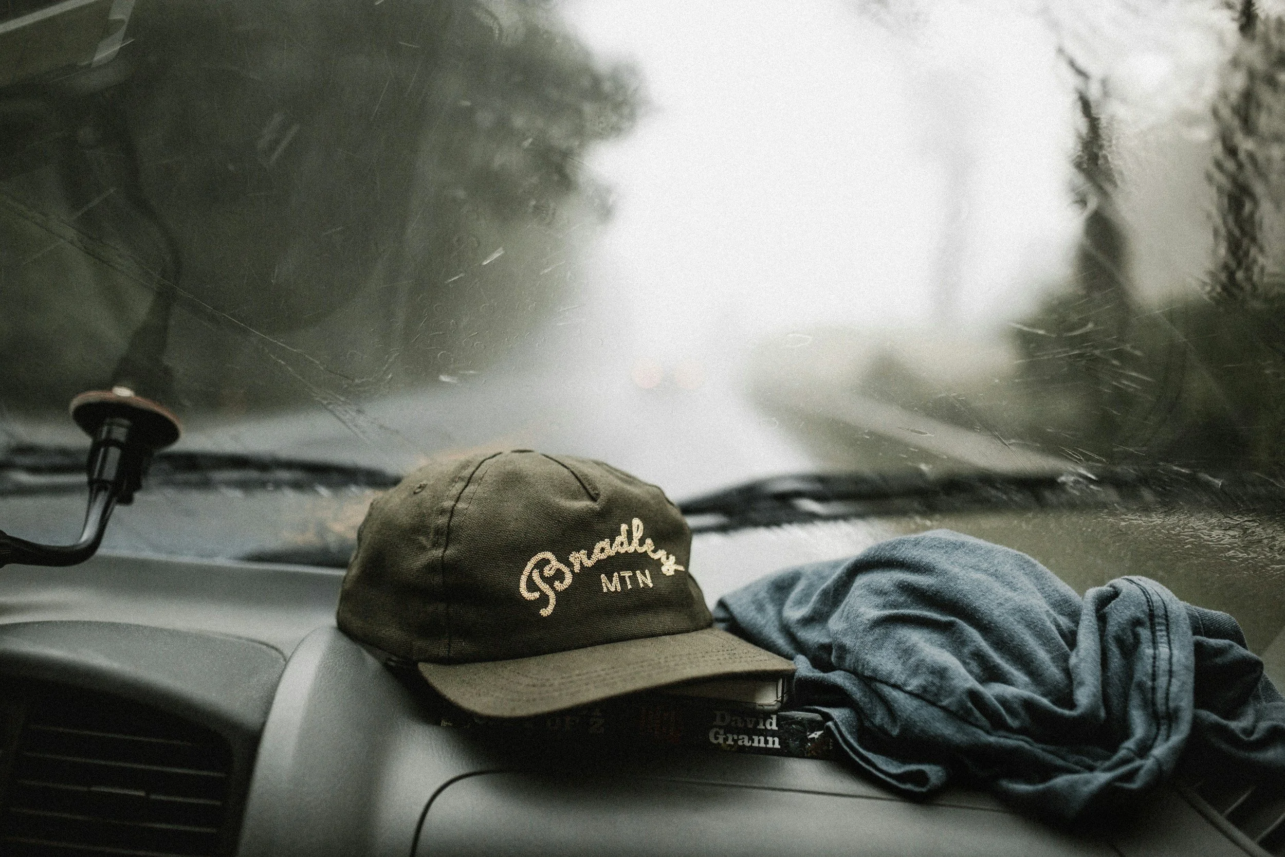 Cappello verde con scritta 'Bradley MTN' e pantaloni di jeans sul cruscotto di un'auto, vista attraverso il parabrezza bagnato dalla pioggia.
