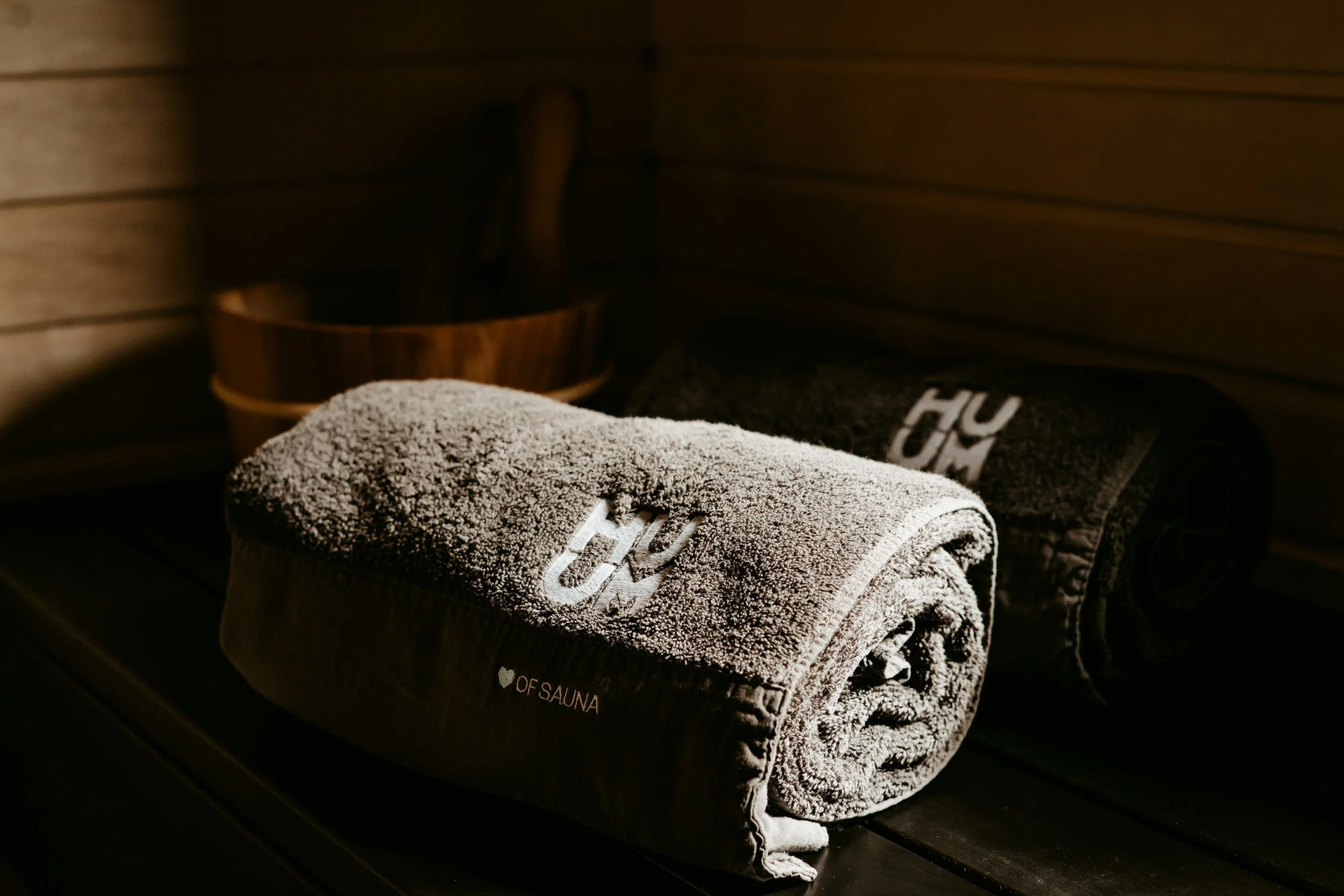 Asciugamani grigi e neri arrotolati in una sauna, con una scritta che indica il cuore of sauna, e in sfondo un secchio di legno con un cucchiaio di legno, tutto in un ambiente in legno.