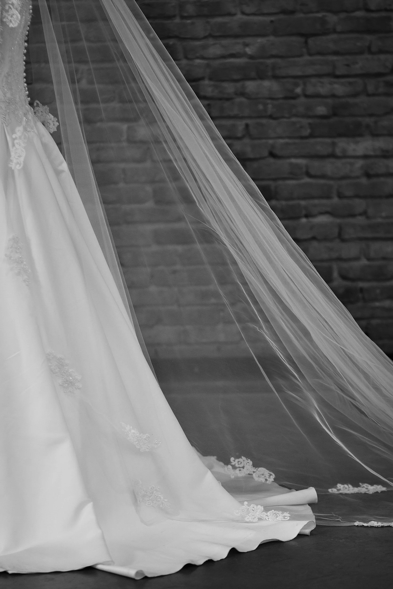 Vestito da sposa bianco con pizzi, in parte coperto da una veletta di tulle trasparente, di fronte a un muro di mattoni.