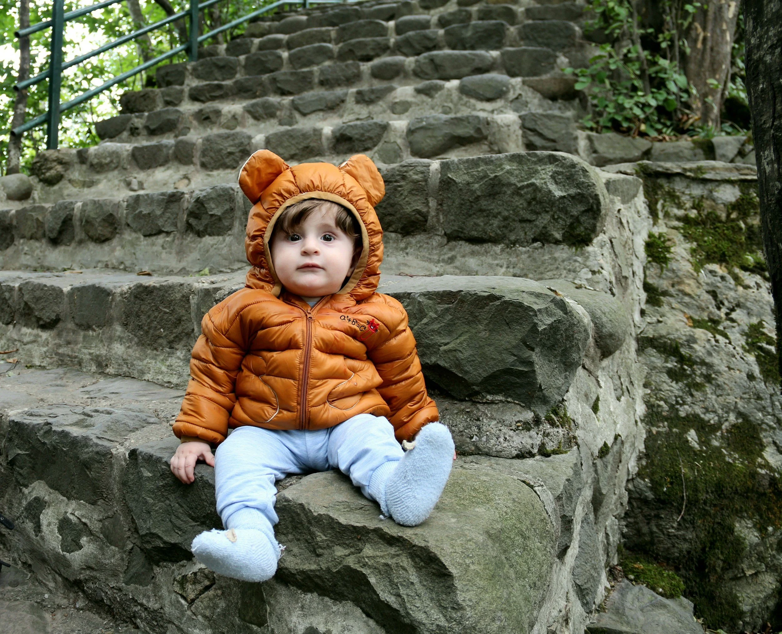 Bambino seduto su scalini in pietra, indossa una giacca marrone con cappuccio a forma di orsetto e calze bianche, ambientato in un ambiente naturale con vegetazione e gradini di pietra.